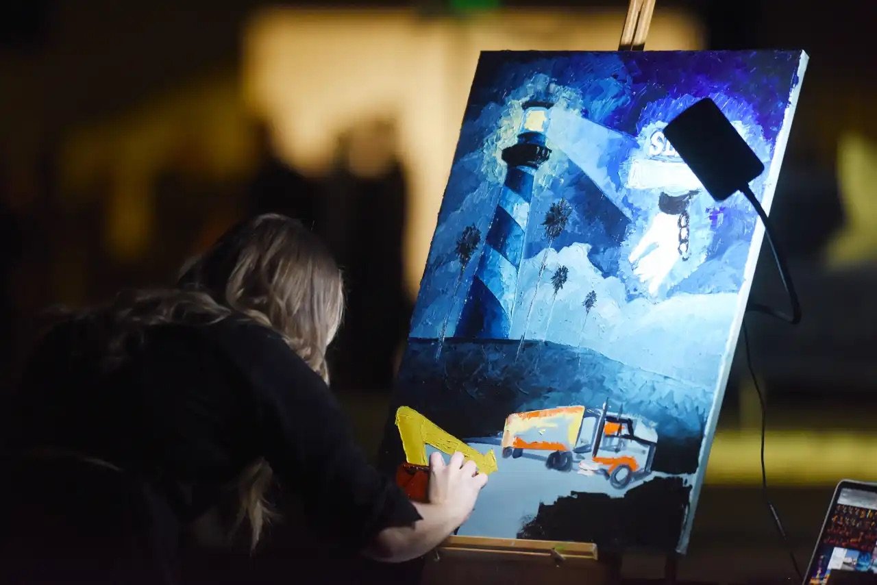 Person painting a landscape scene of a lighthouse, palm trees, and a truck at night on a canvas on an easel.