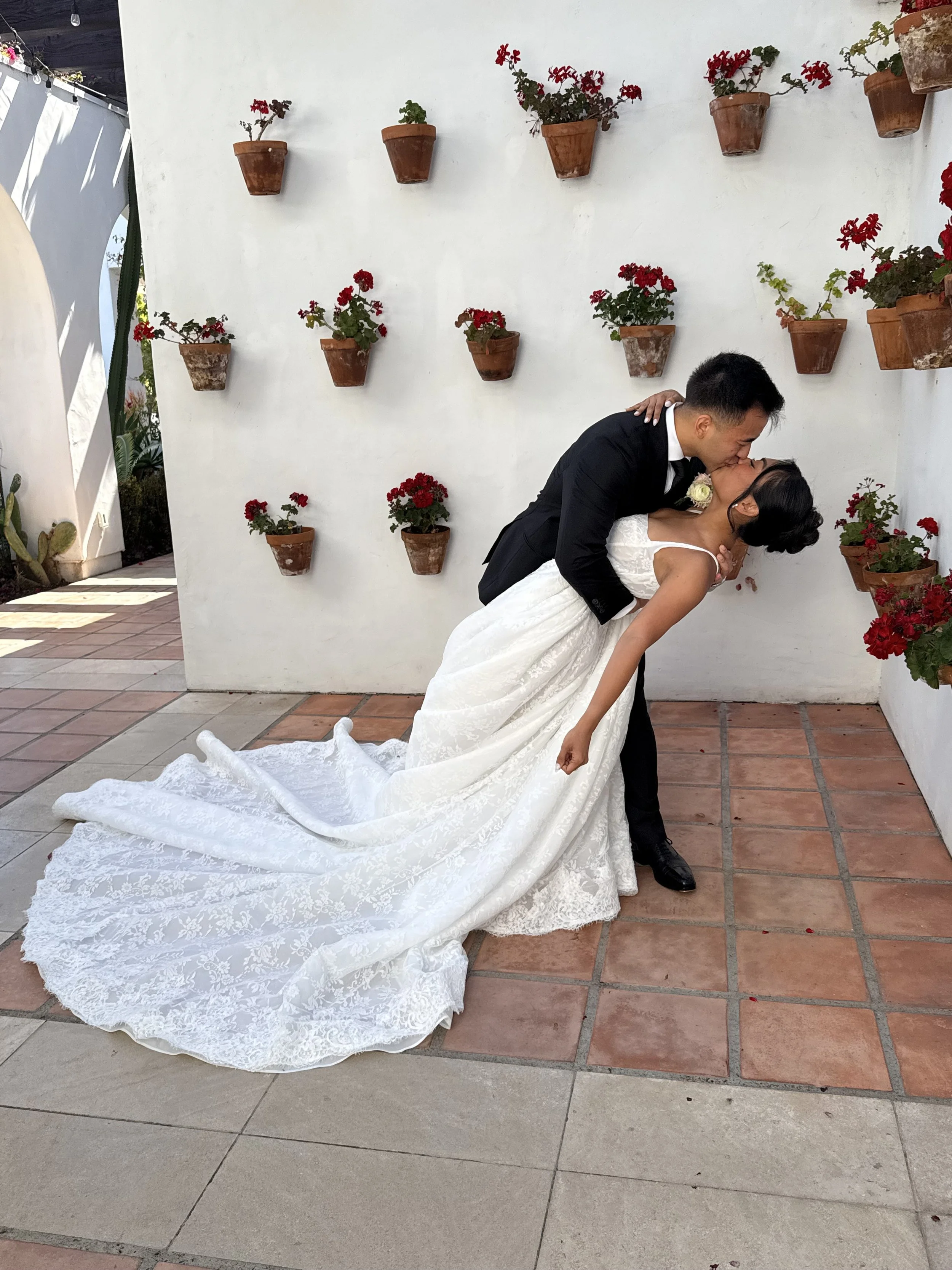 A newlywed couple kissing, with the groom dipping the bride, in front of a white wall decorated with potted red flowers.