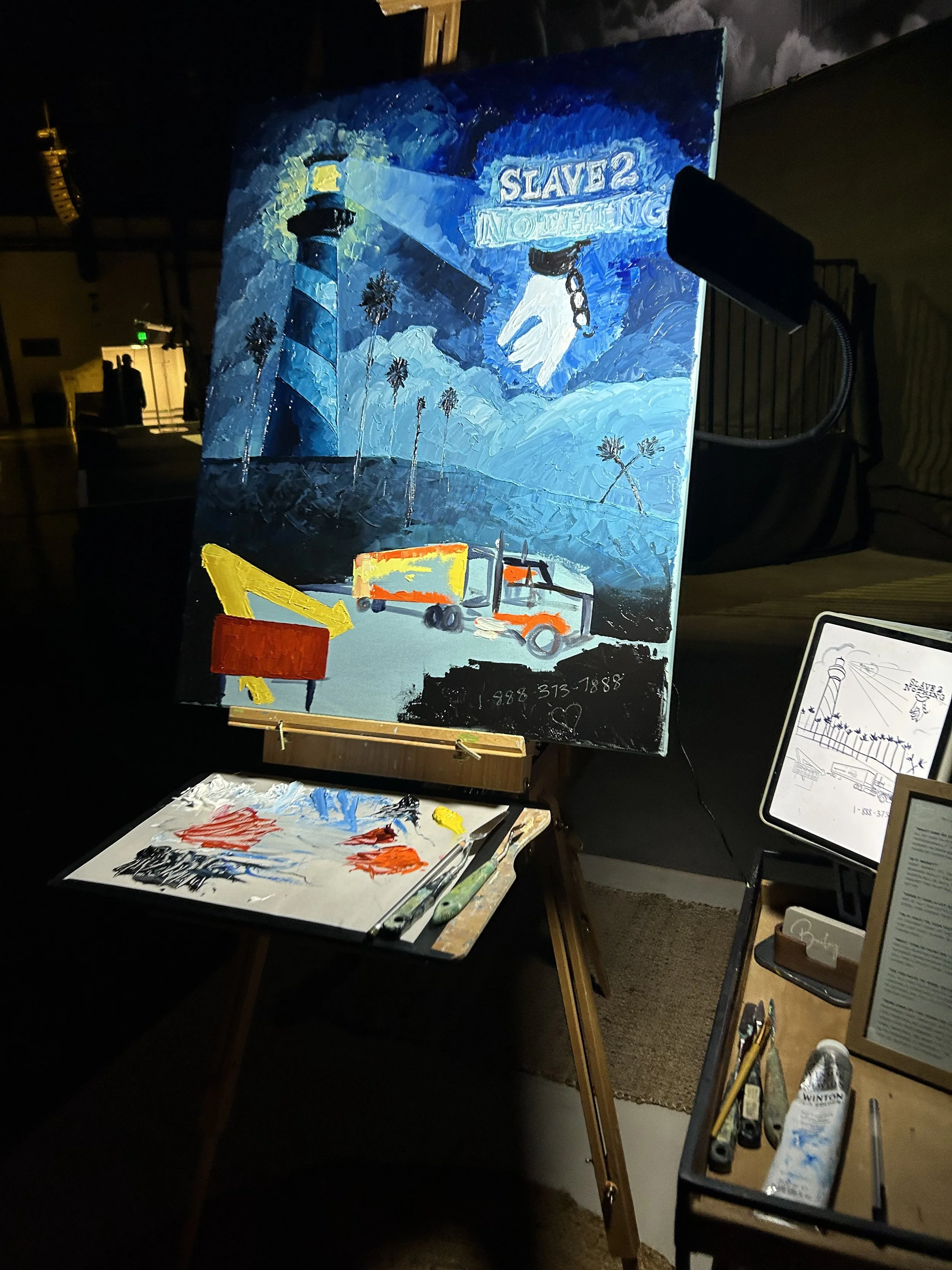 Painting depicts a lighthouse at night with a beam of light shining out, surrounded by mountains and trees. Text on the painting reads 'SLAVE 2 NOTHING.' Below the painting, there is a palette with mixed paints, and a small orange and red object, possibly a toy or model. The setting appears to be an artist's studio with various art supplies and sketches visible.