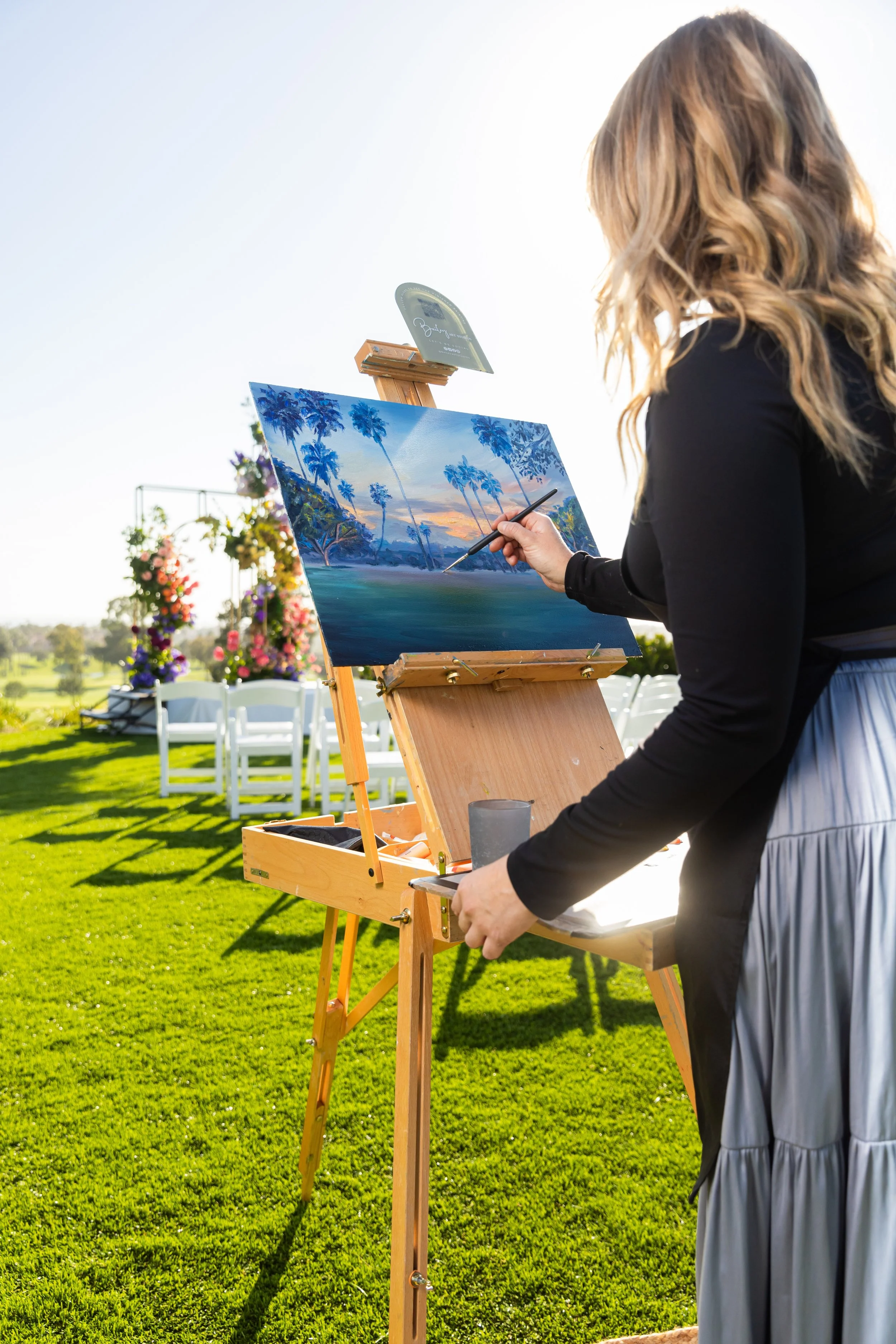 A woman painting a landscape scene on an outdoor canvas set up on an easel during the daytime. The painting depicts palm trees at sunset. The woman has long, wavy blonde hair and is dressed in a black top and a gray skirt. In the background, there are white chairs and flower arrangements, suggesting a garden or outdoor event setting.