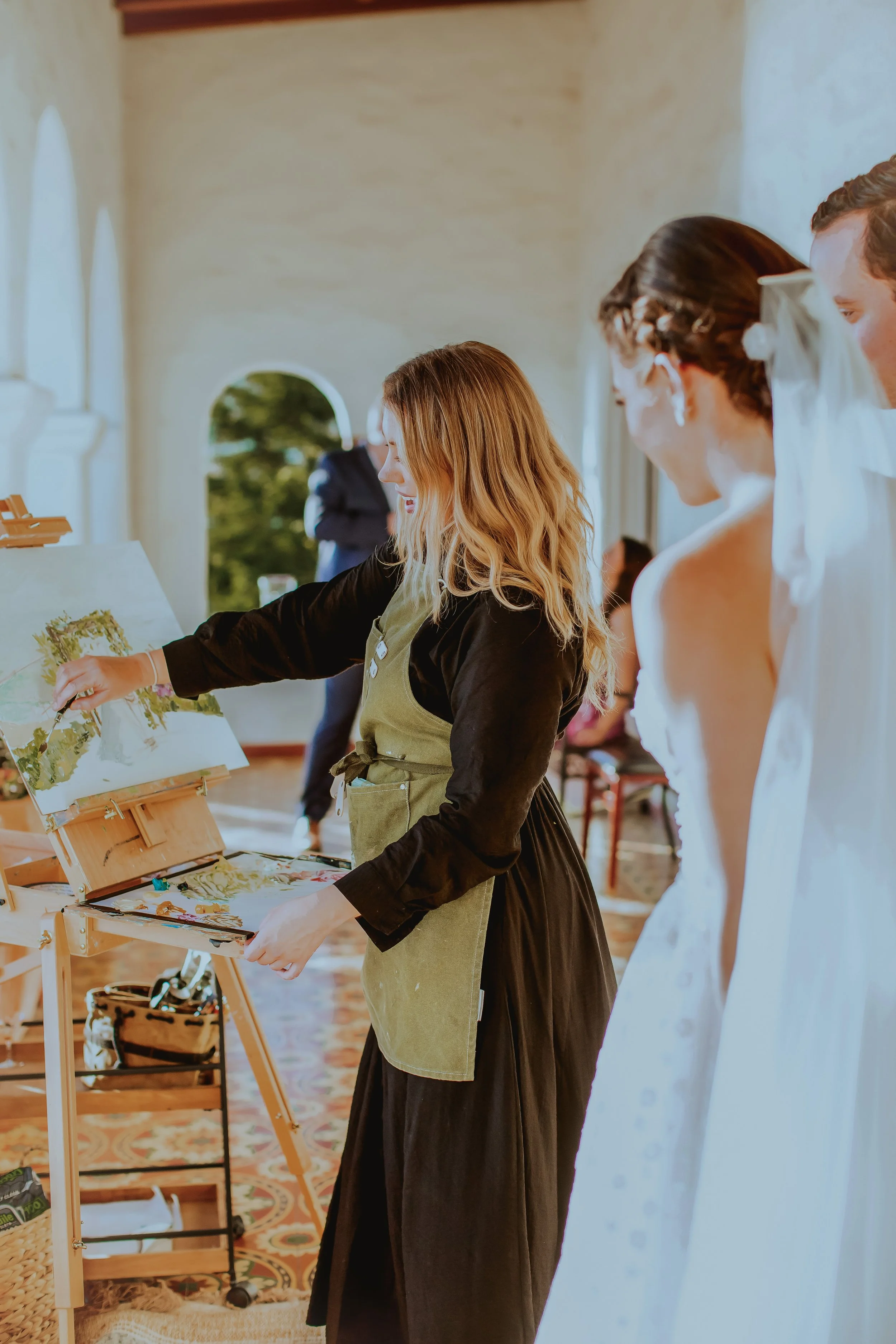 People gathered at a daytime wedding, with women in white dresses and a woman painting on a canvas.