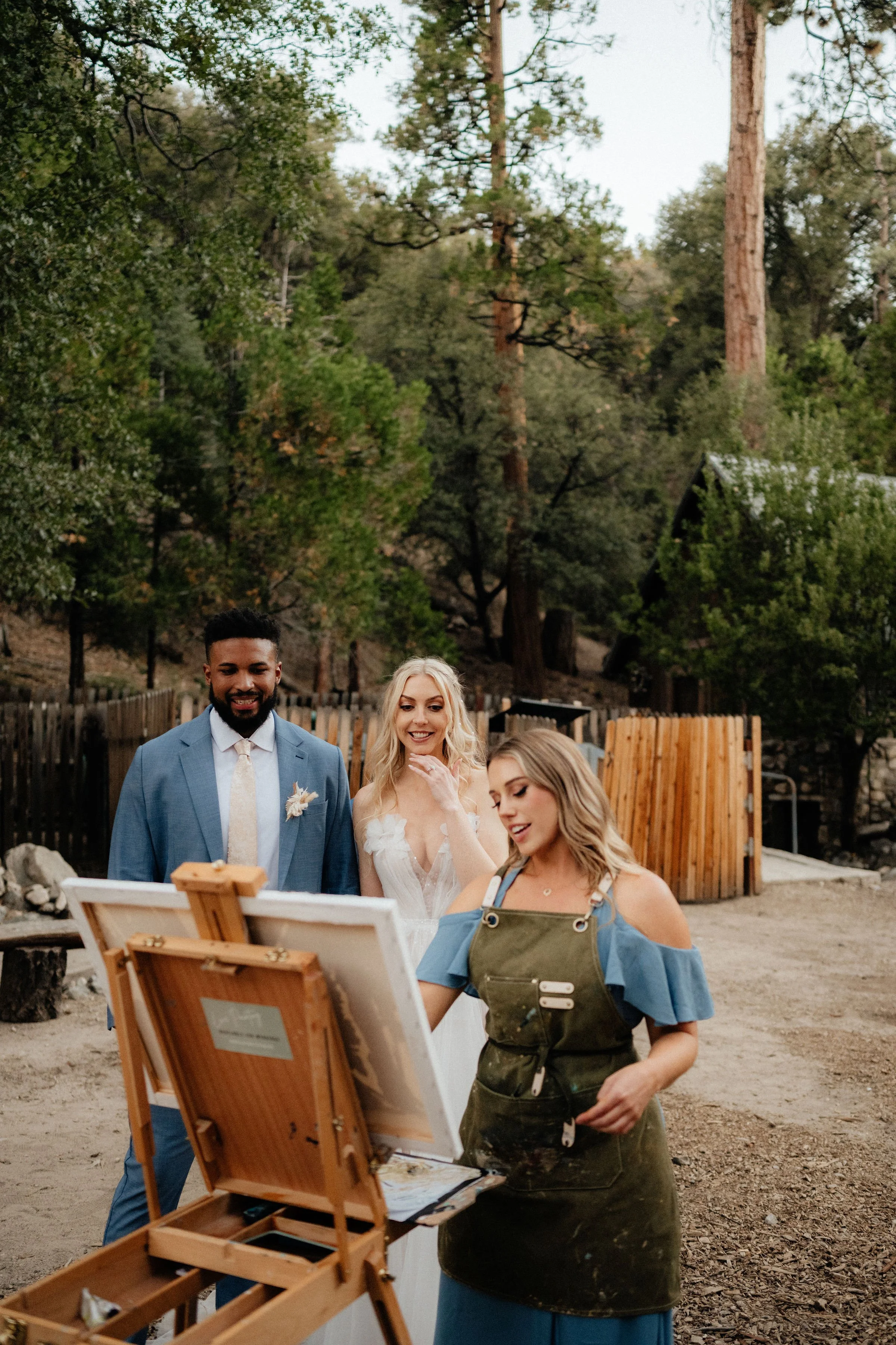 A group of people participating in an outdoor painting activity at a rustic location with trees and wooden fences, with a woman in an apron actively painting on a canvas.