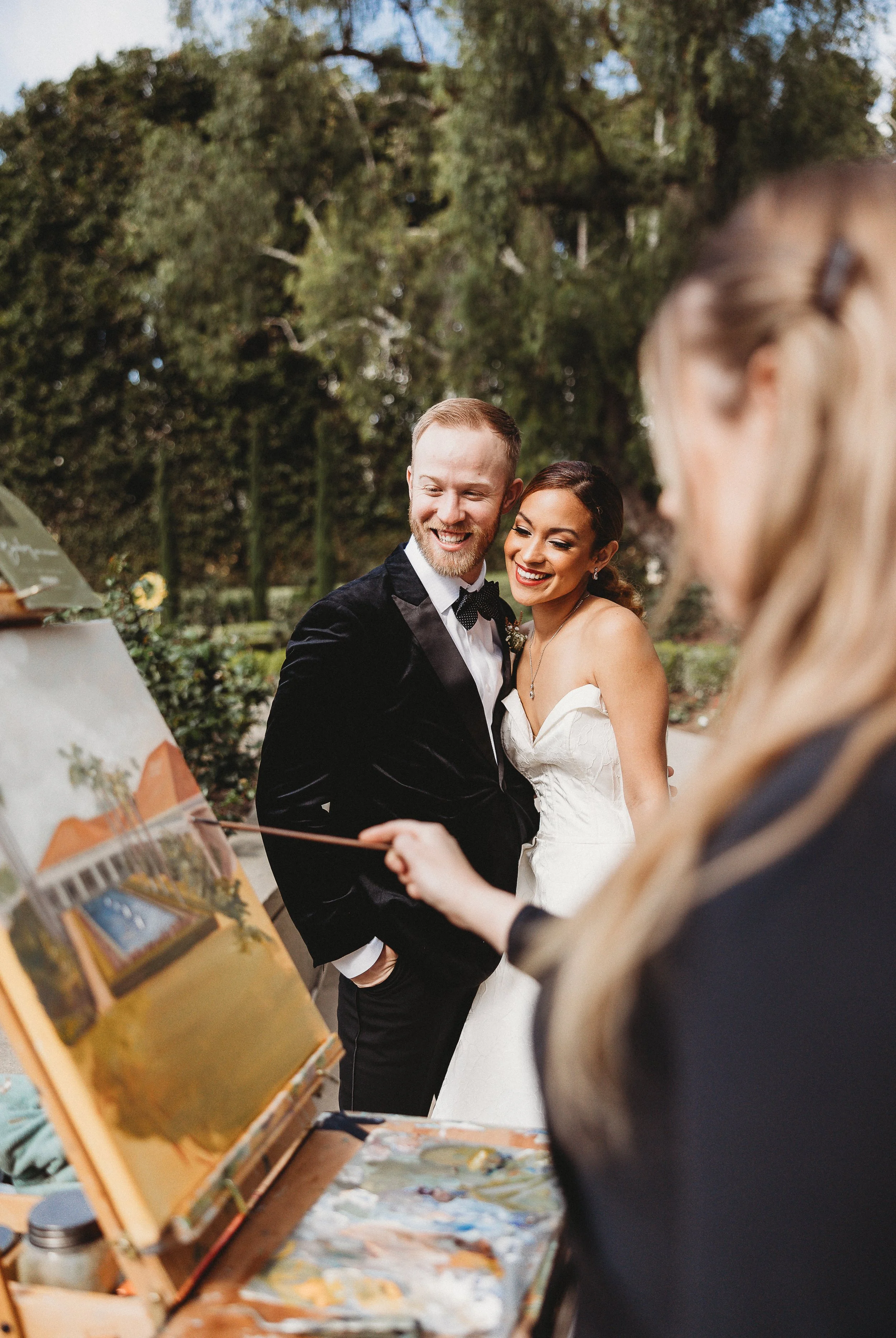 A bride and groom smiling at an outdoor wedding ceremony. The groom wears a tuxedo, and the bride wears a wedding gown. An artist is painting a landscape on an easel nearby.