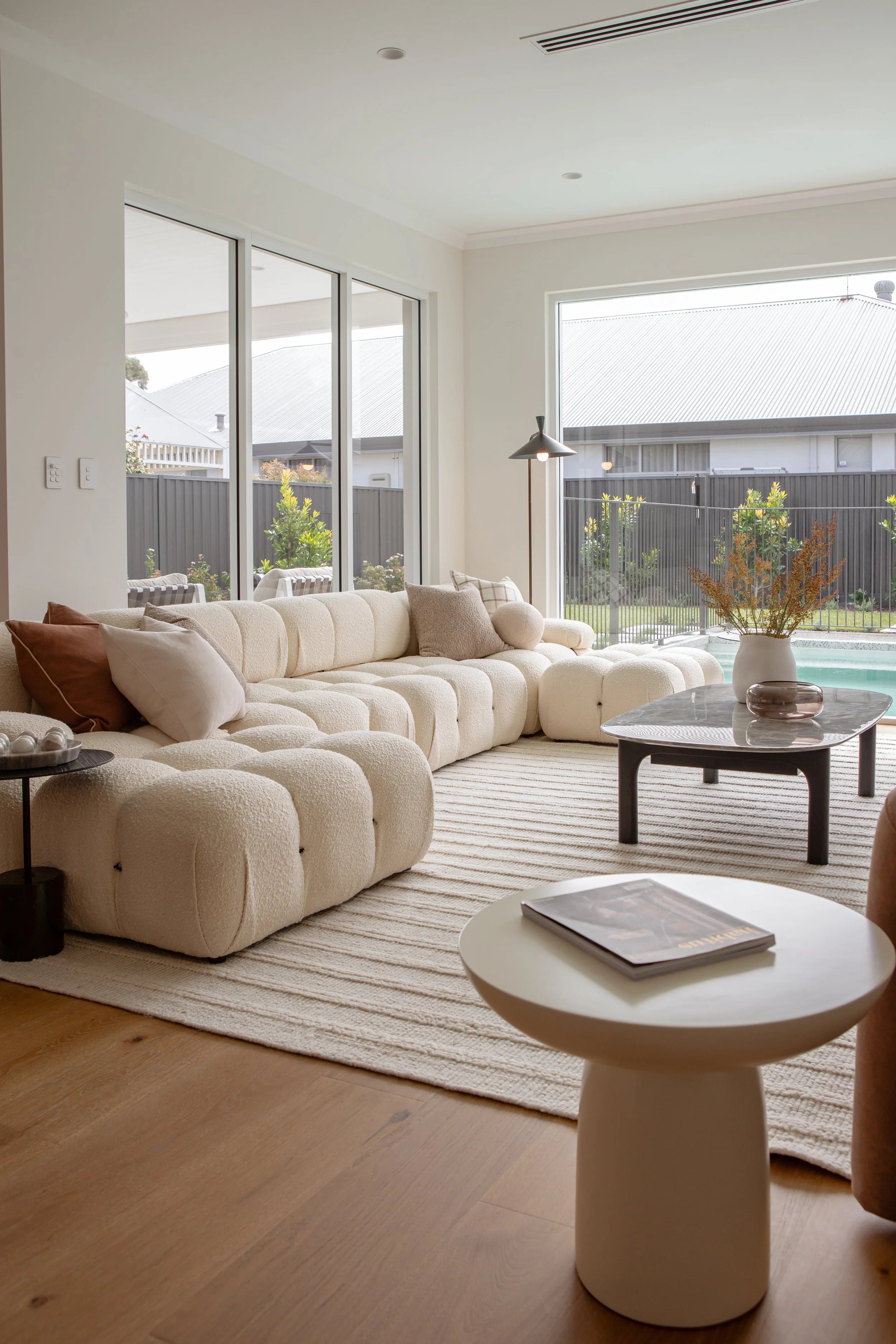 Living room with a large, cream-colored, tufted sectional sofa, cushions, a striped rug, a small side table with a book, a coffee table with decorative items, and large windows showing a backyard with a fence, trees, and a pool.