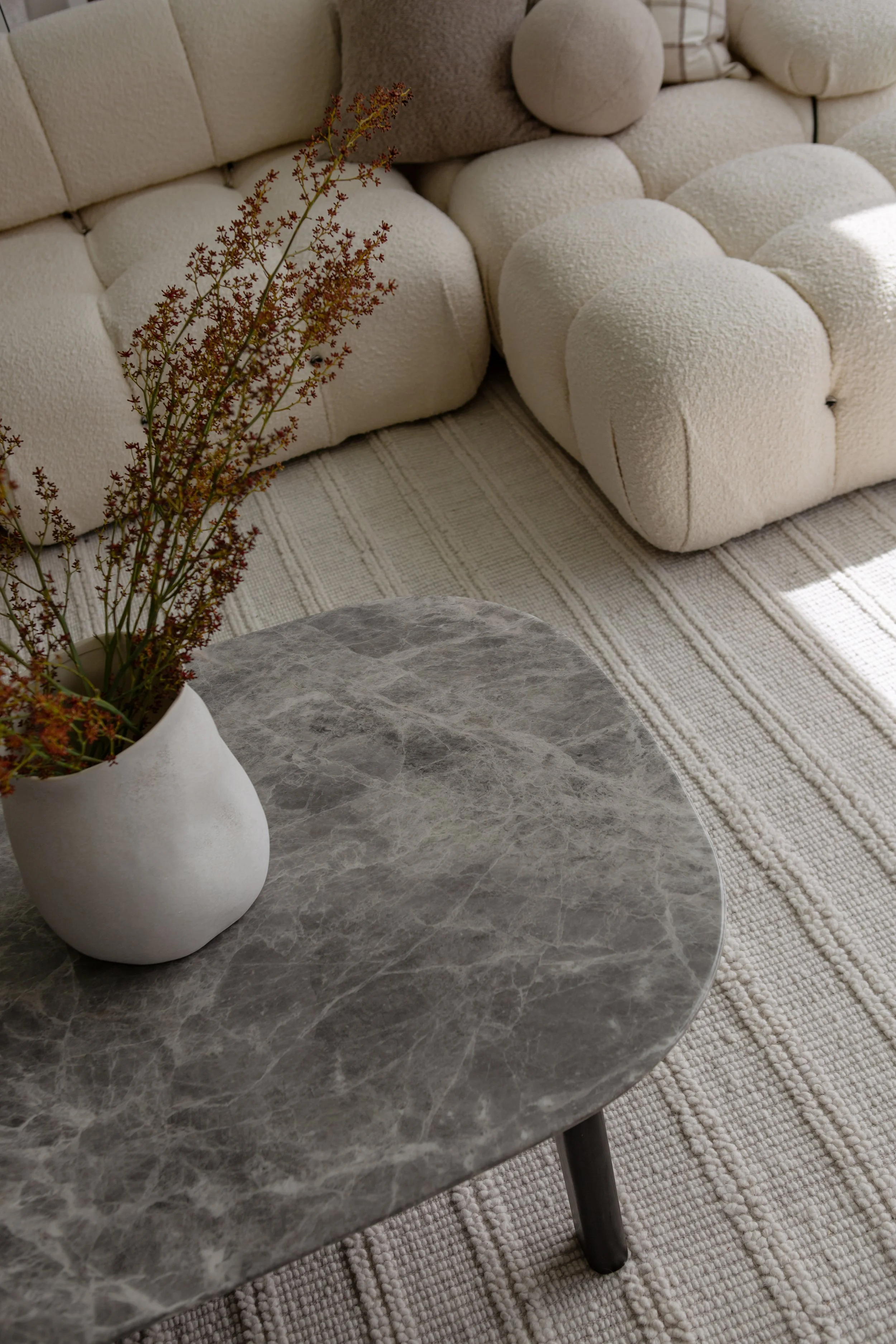 A living room with a textured beige sofa, a round gray marble coffee table, and a white ceramic vase with dried flowers, on a striped beige rug.