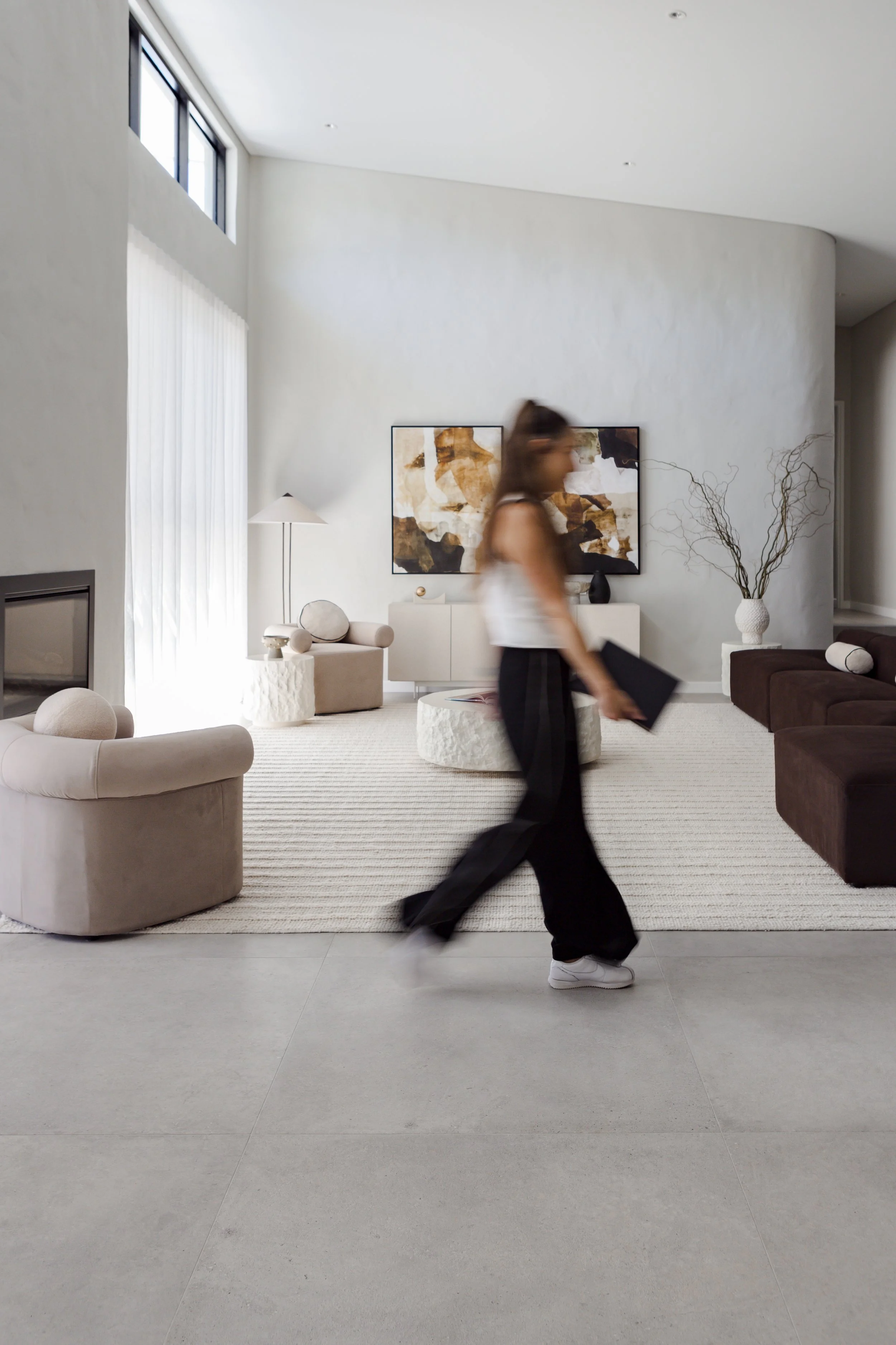 Blurry woman walking through a modern, minimalist living room with white and beige furnishings, a dark brown sofa, and abstract artwork on the wall.