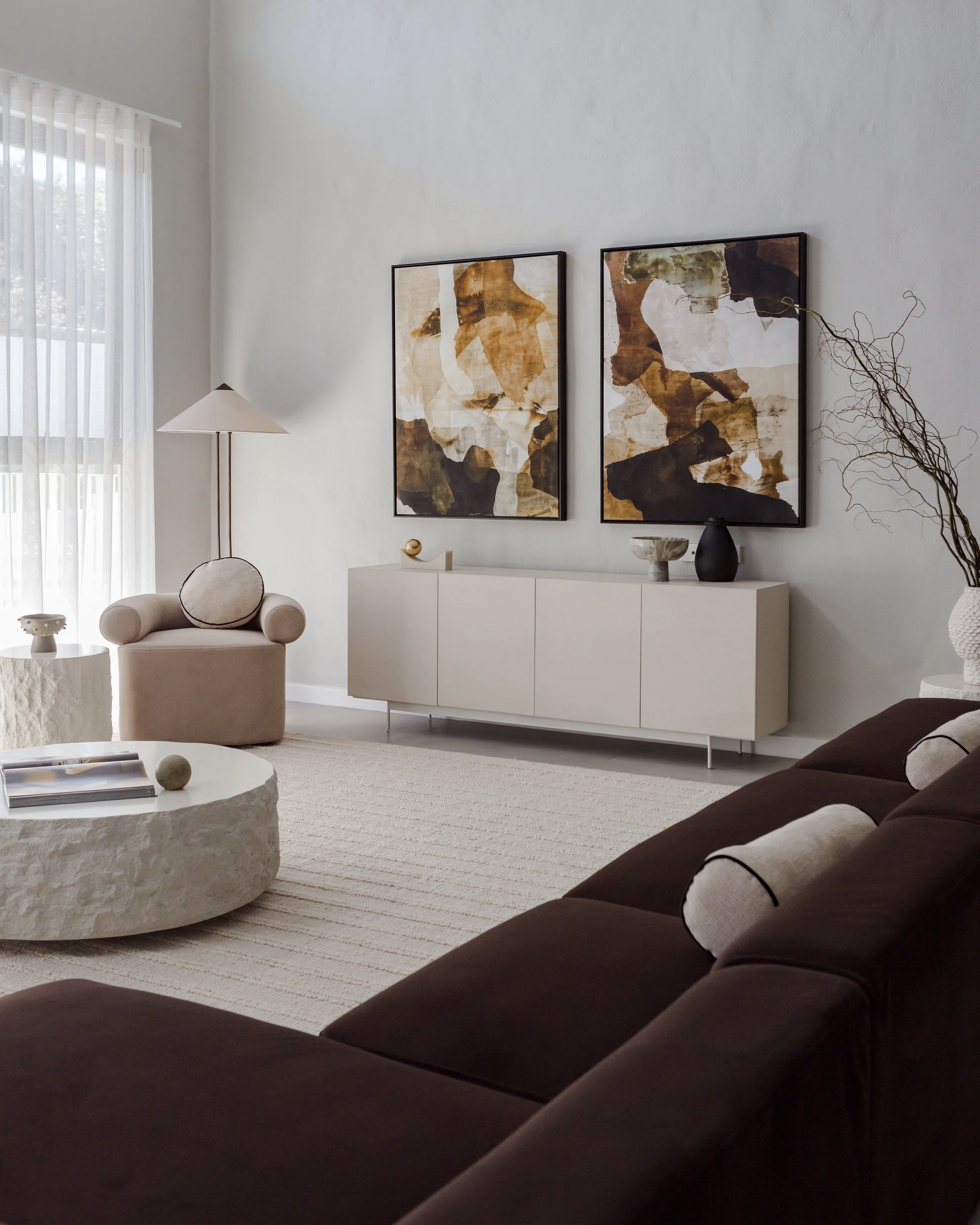 Modern living room with beige armchair, dark brown sofa with pillows, white textured coffee table, large window with sheer white curtains, artwork on the wall, and decorative vases.