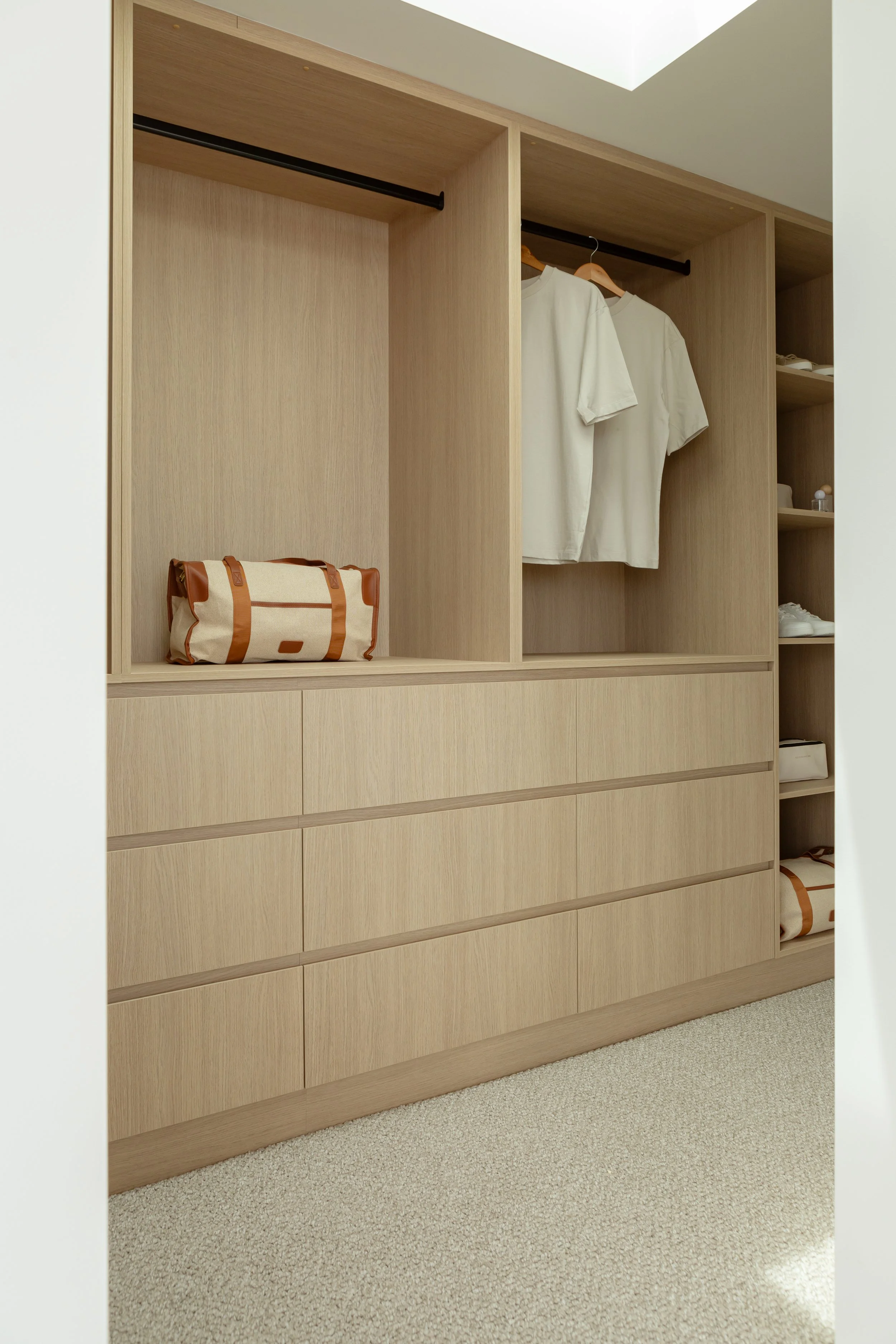 A built-in wooden wardrobe with open compartments, containing two white shirts on hangers, a beige and brown duffle bag, and some folded clothing on the shelves. The wardrobe has multiple drawers at the bottom and is set against a light wall with a ceiling light above.