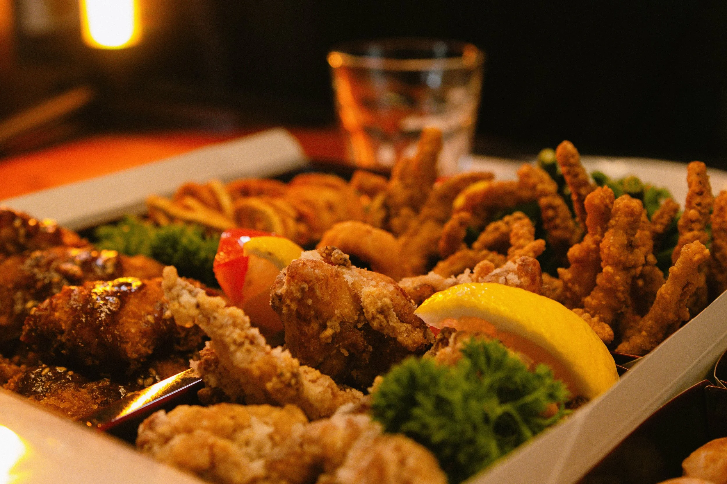 Fried chicken wings, lemon slices, broccoli, and other fried appetizers on a festive dinner table with a glass of water and blurred candle in the background. 奥克兰武藏日本料理餐厅. Musashi Japanese Restaurant, Auckland, St Heliers & Milford.