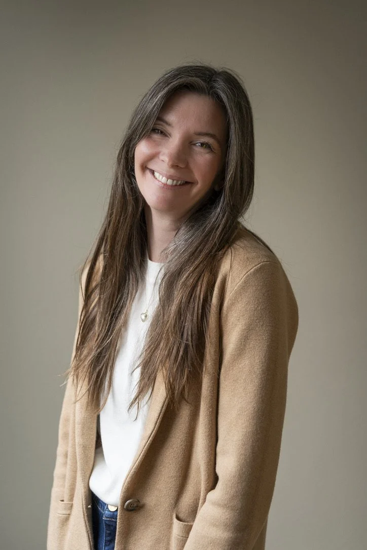 A woman with long brown hair smiling in a casual beige blazer and white shirt against a neutral background.