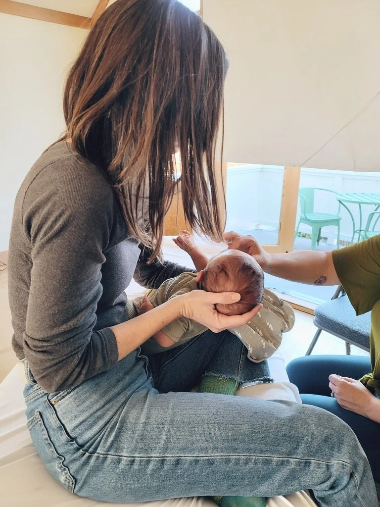 A woman with brown hair is holding a newborn baby on her lap while another person, wearing a green shirt, gently touches the baby's head.