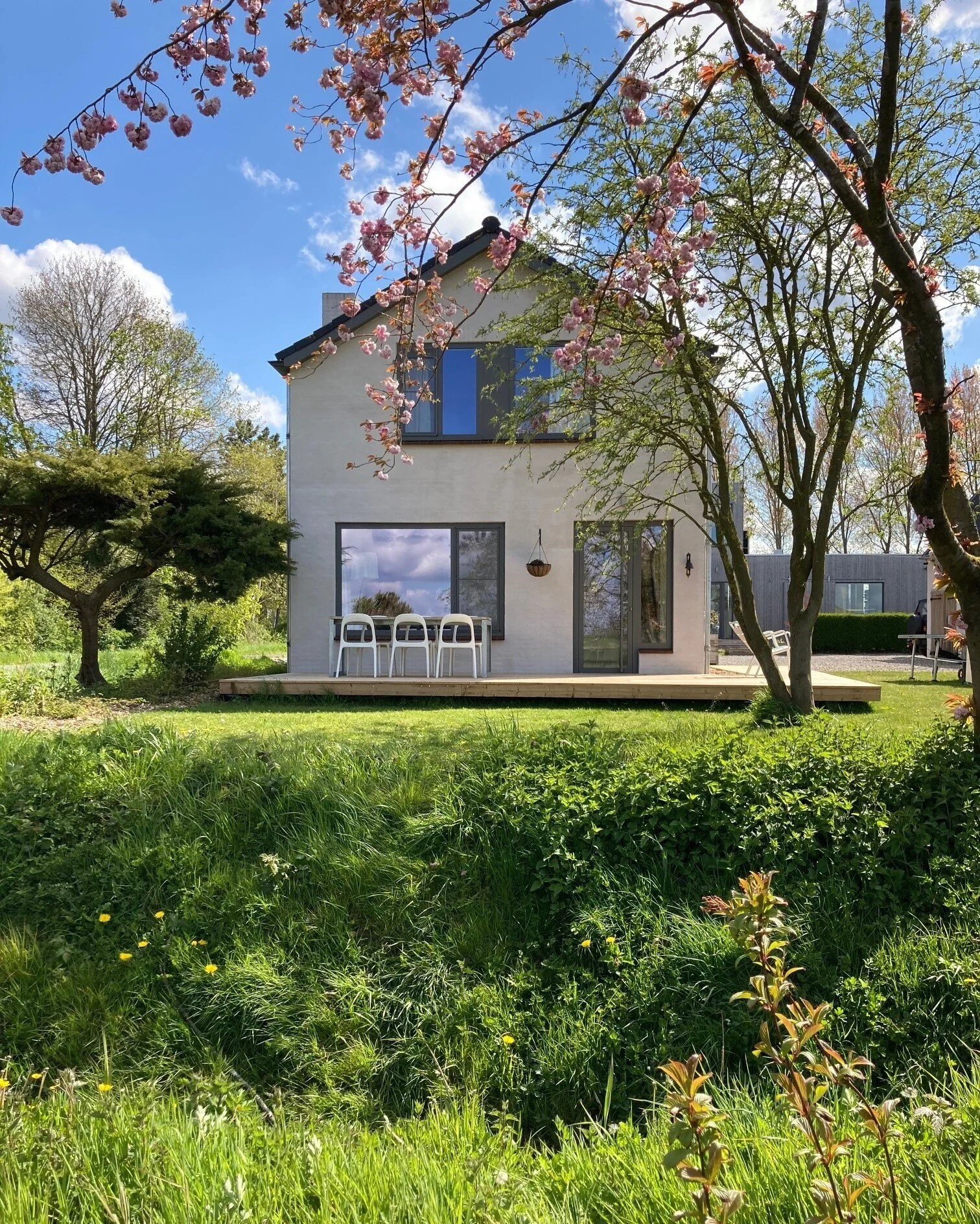 Moderne witte huiskamer met grote ramen, uitzicht op tuin en tuinmeubilair, met groene planten.