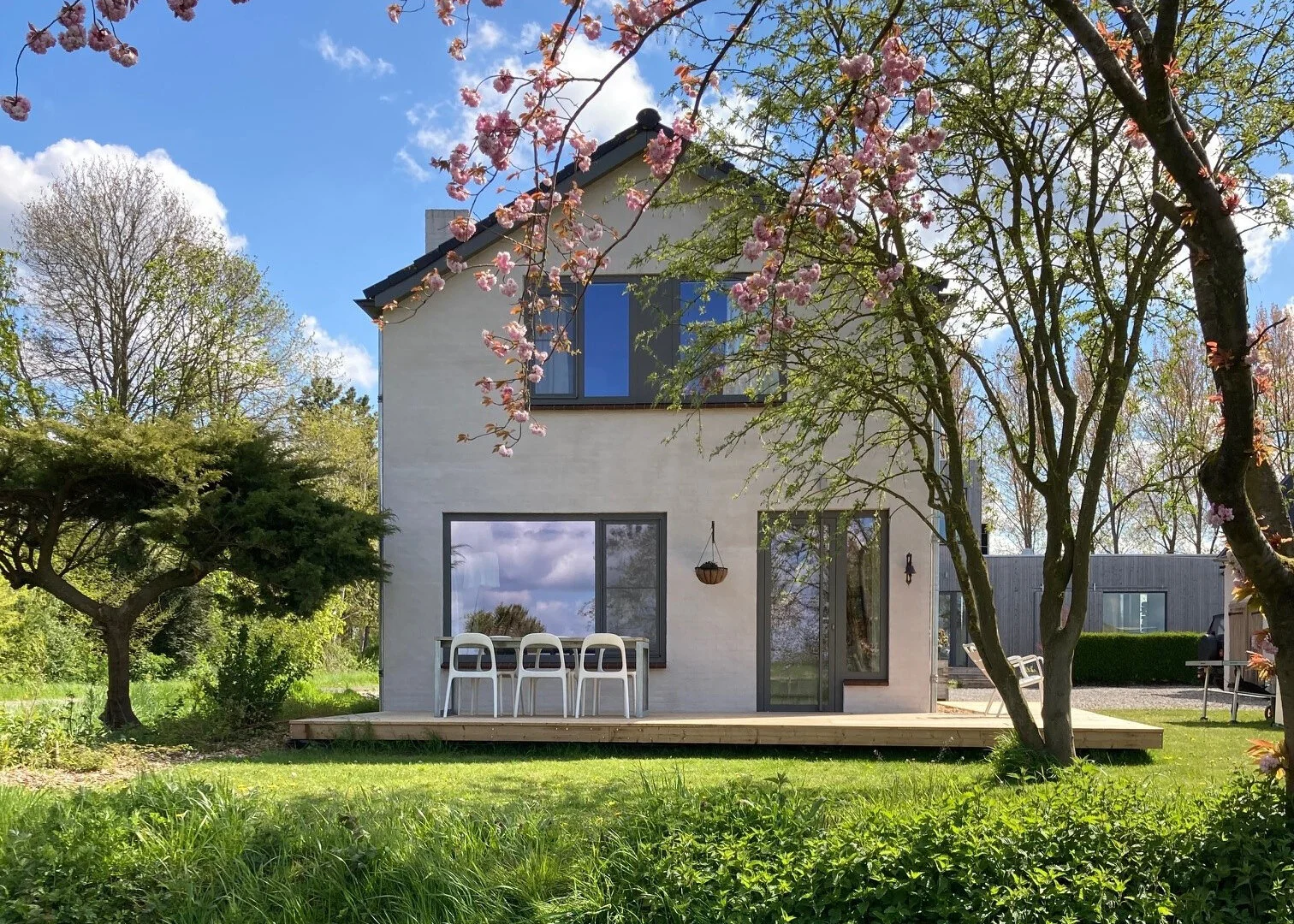 Moderne witte twee-verdiepingen huis met grote ramen, terras met stoelen, omgeven door groene bomen en bloemen, onder een blauwe lucht met witte wolken.