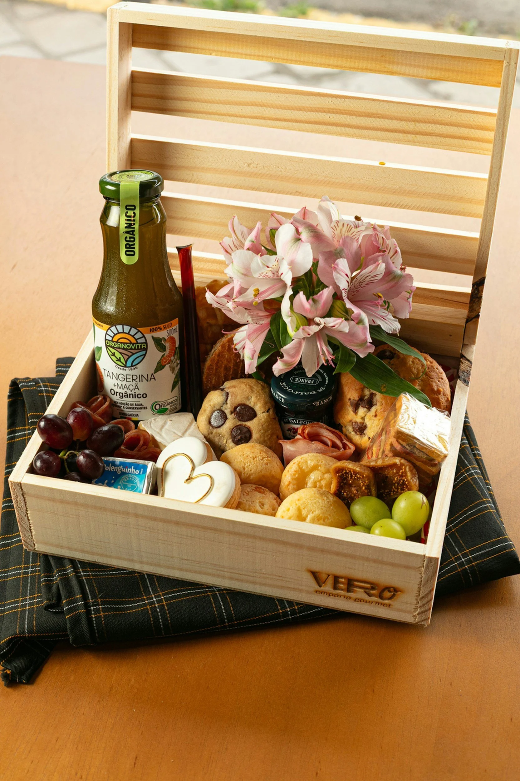 Prijzenbox gevuld met diverse snacks, koekjes, fruit, bloemen en een fles sap, geplaatst op een tafel met een zwart-wit geruite tafelkleed.
