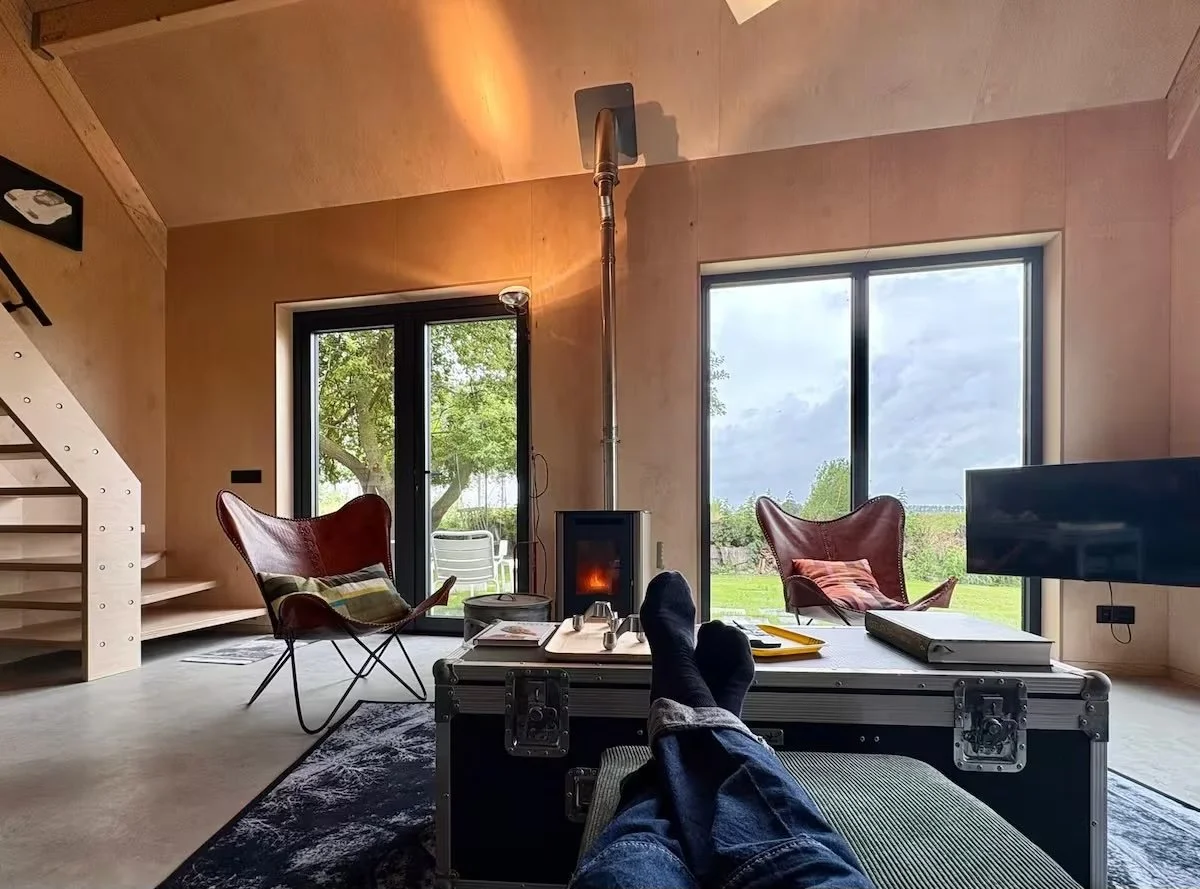 Binnenkant van De Studio, een vakantiewoning te huur met woonkamer met grote ramen die uitzicht bieden op de tuin, een houtkachel, twee rode loungestoelen en een televisie.