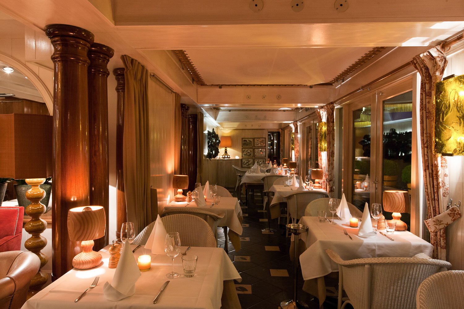 Een restaurant met gedekte tafels, kaarsen, glazen en servetten, sfeervolle verlichting en grote ramen met uitzicht op de buitenlucht 's avonds.
