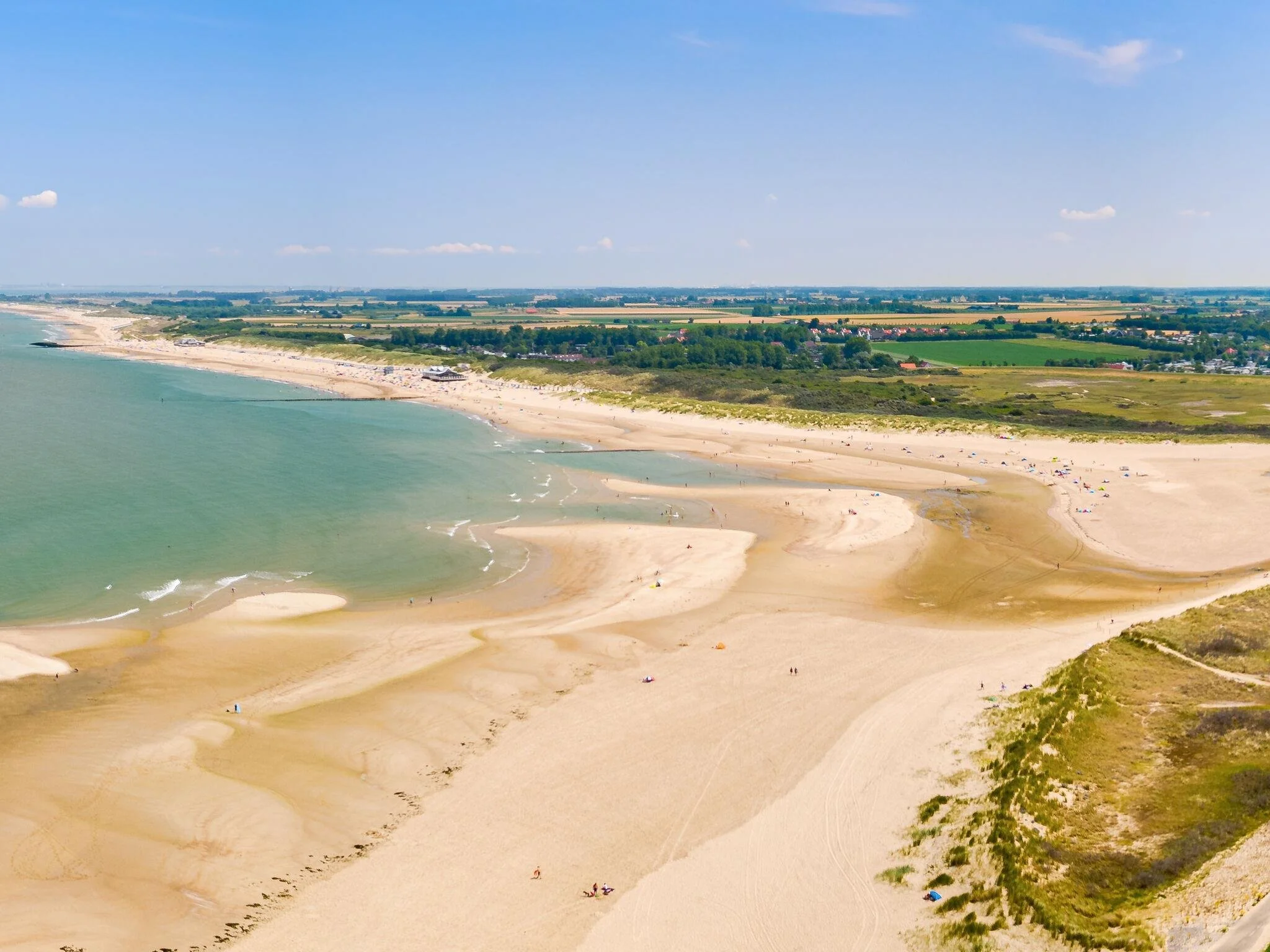 Uitzicht op een breed zandstrand langs de kust met mensen die ervan genieten, groene weilanden en bossen in de achtergrond onder een heldere blauwe lucht.