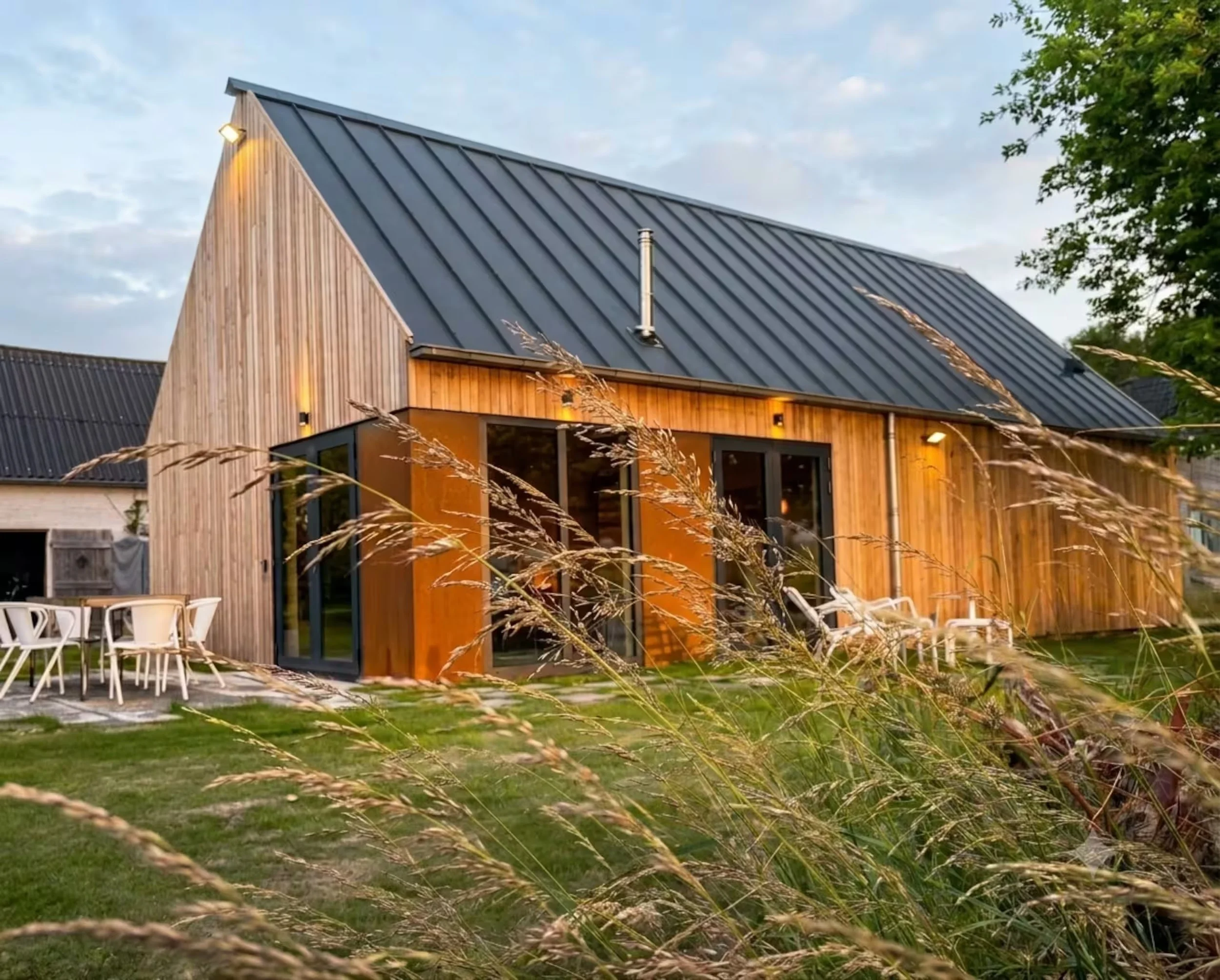 De Dijck Studio, moderne houten huis huren in Zeeland met grote ramen en een metaal dak, omgeven door gras en bomen, 's avonds met verlichting