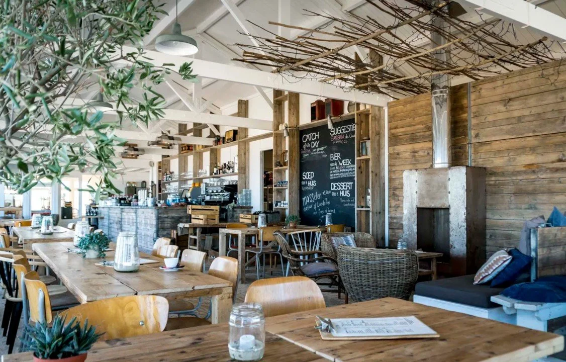 Interieur van restaurant Loods 10 in Breskens met houten tafels en stoelen, groene planten en een open haard, met natuurlijke lichtelementen en decoratie.