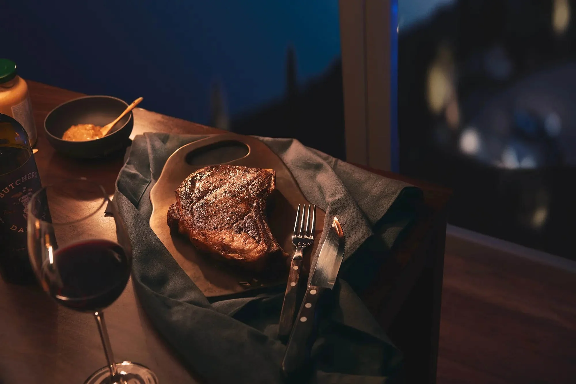 Ein Tisch mit einem Serviertablett und einem saftigen, gebratenen Steak. Daneben liegt Besteck, ein Glas Rotwein und eine Schale mit Senf oder Sauce. Die Szene wirkt gemütlich bei gedämpftem Licht.