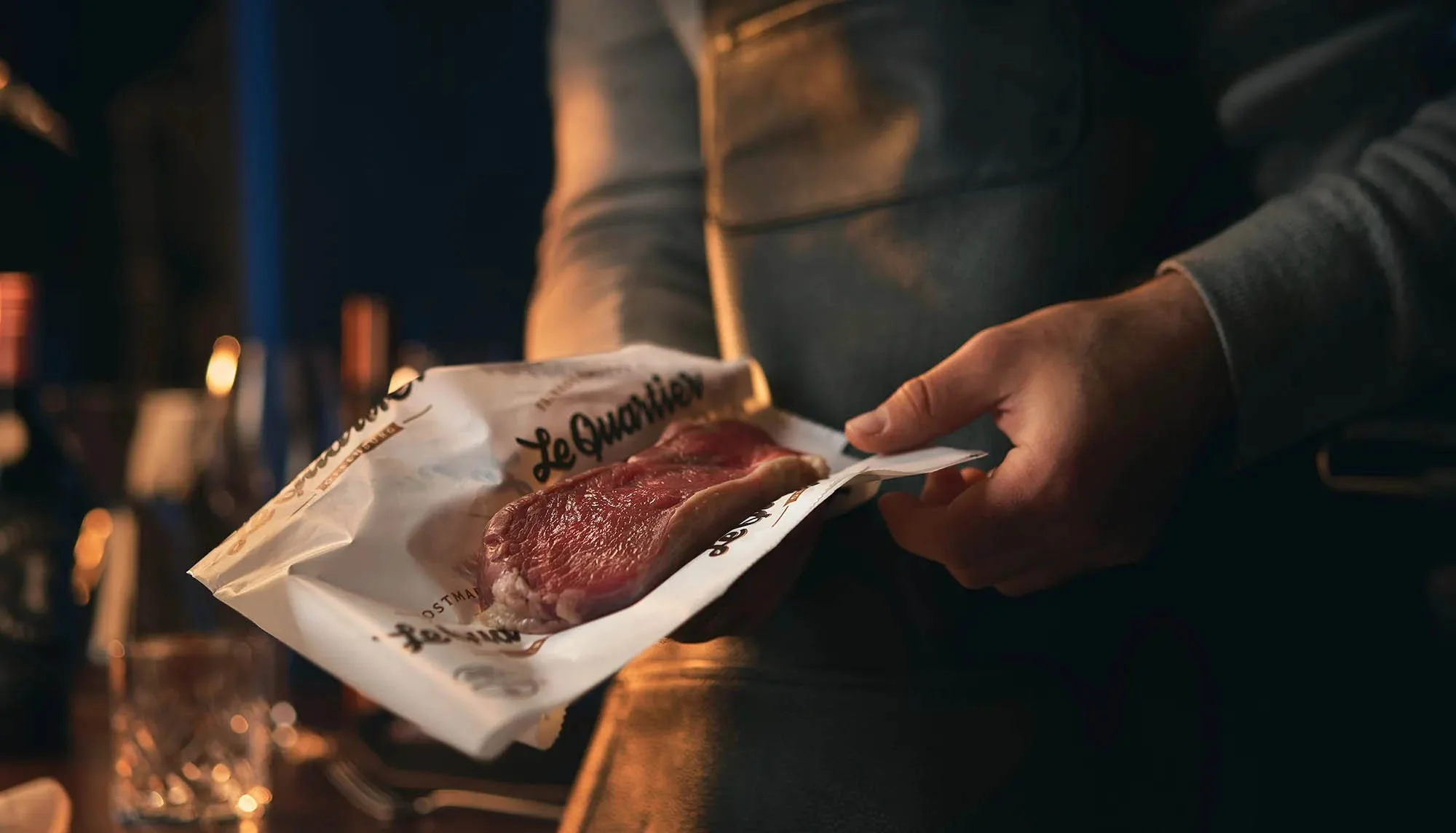 Person hält ein frisches Stück Fleisch in Papierverpackung, mit der Aufschrift "Le Quartier Feinkost".