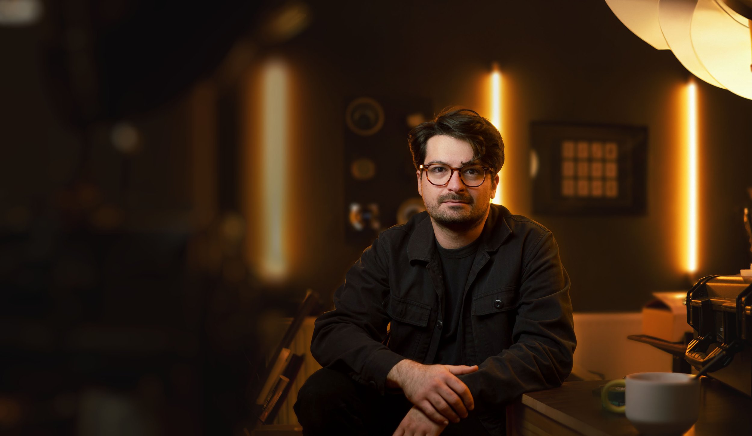 Ein junger Mann mit dunklem Haar, Brille und dunkler Kleidung sitzt in einem dunklen Raum mit warmem Licht im Hintergrund.