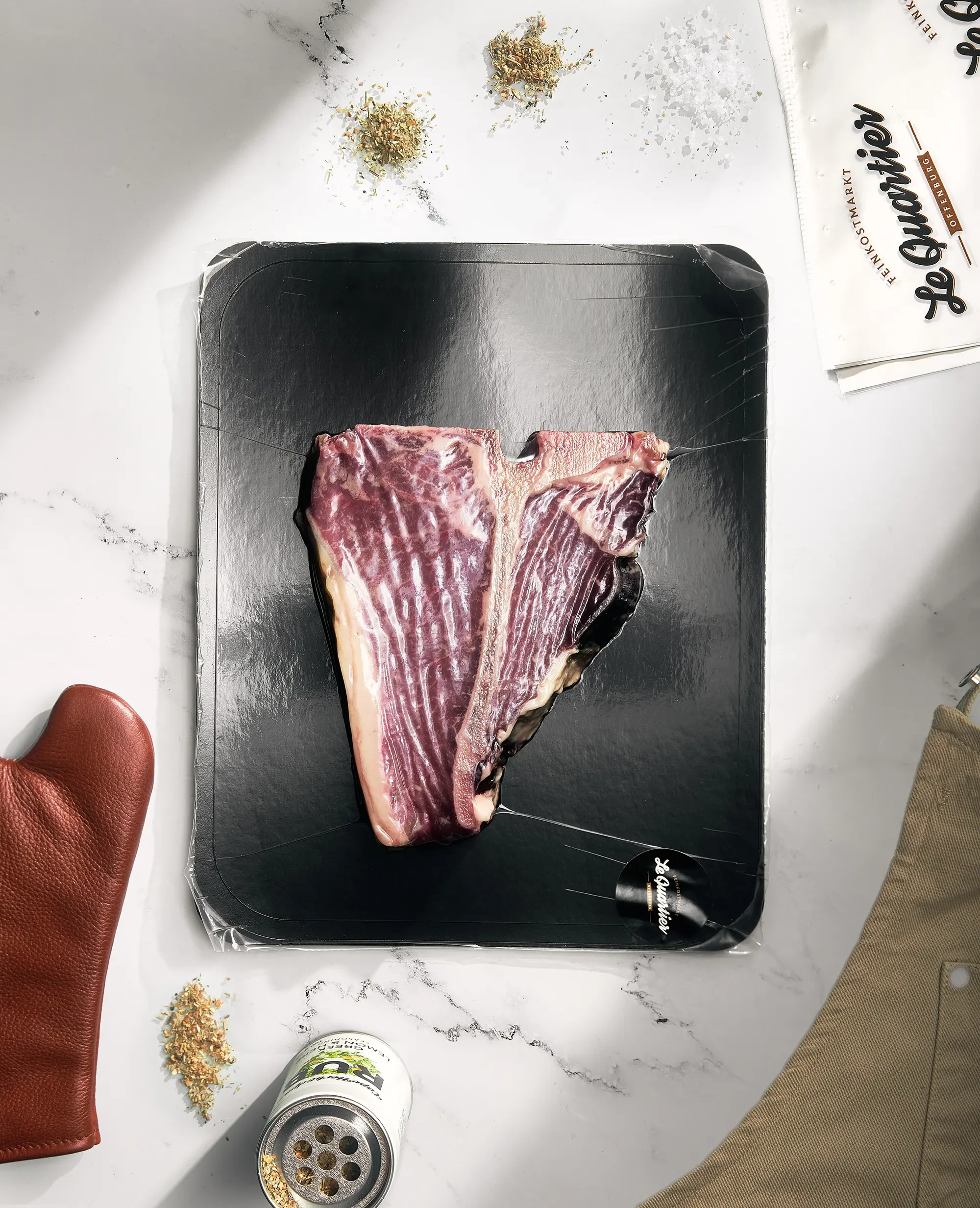 Roher T-Bone Steak auf schwarzem Teller, umgeben von Gewürzen, einer braunen Lederhandschuh, einer Dose und Paprikastreifen auf einem weißen Marmortisch.