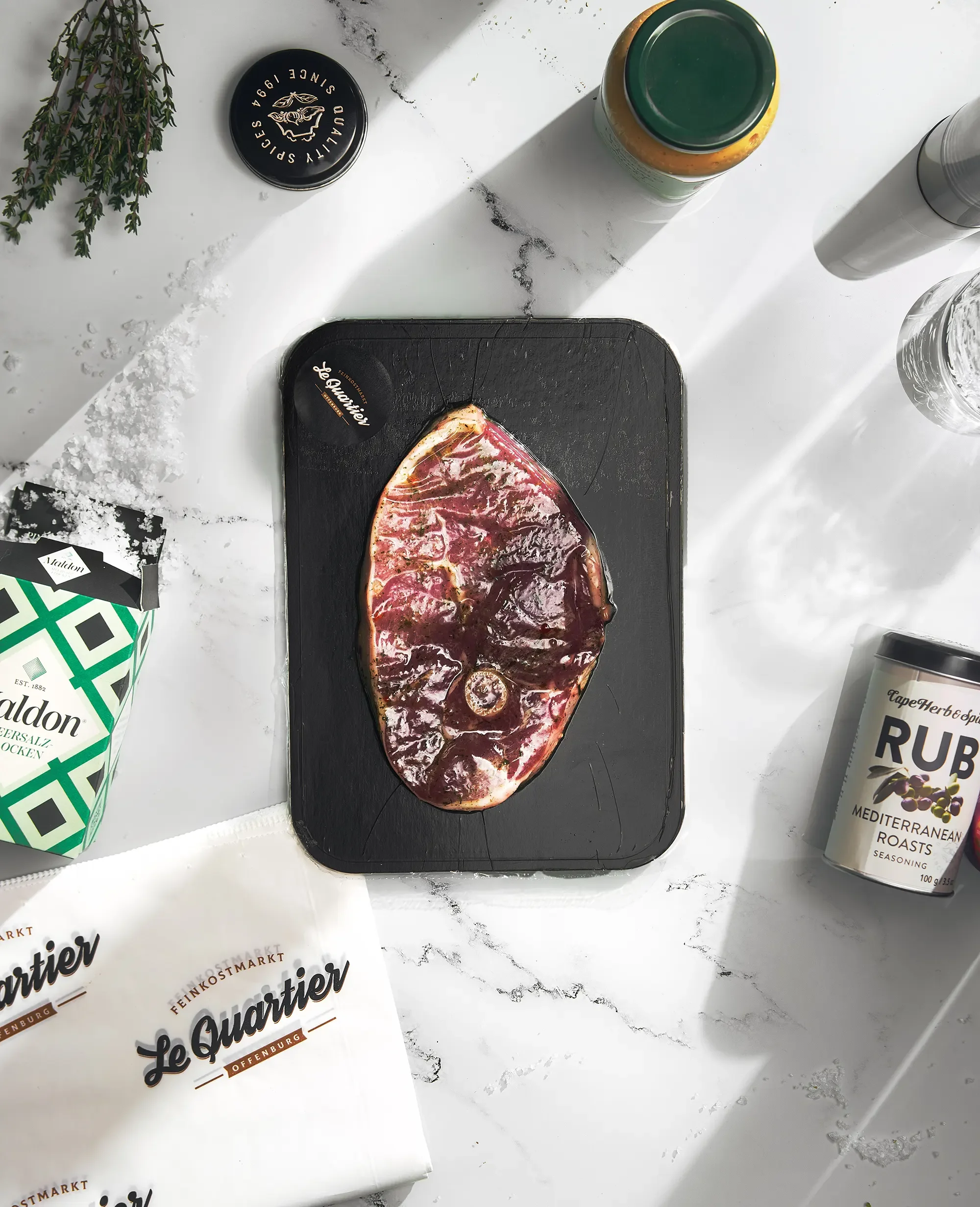 Rindersteak auf einer schwarzen Platte mit Gewürzen, umgeben von Salz, Kräutern, Gewürzbehältern und einem Papiertuch.