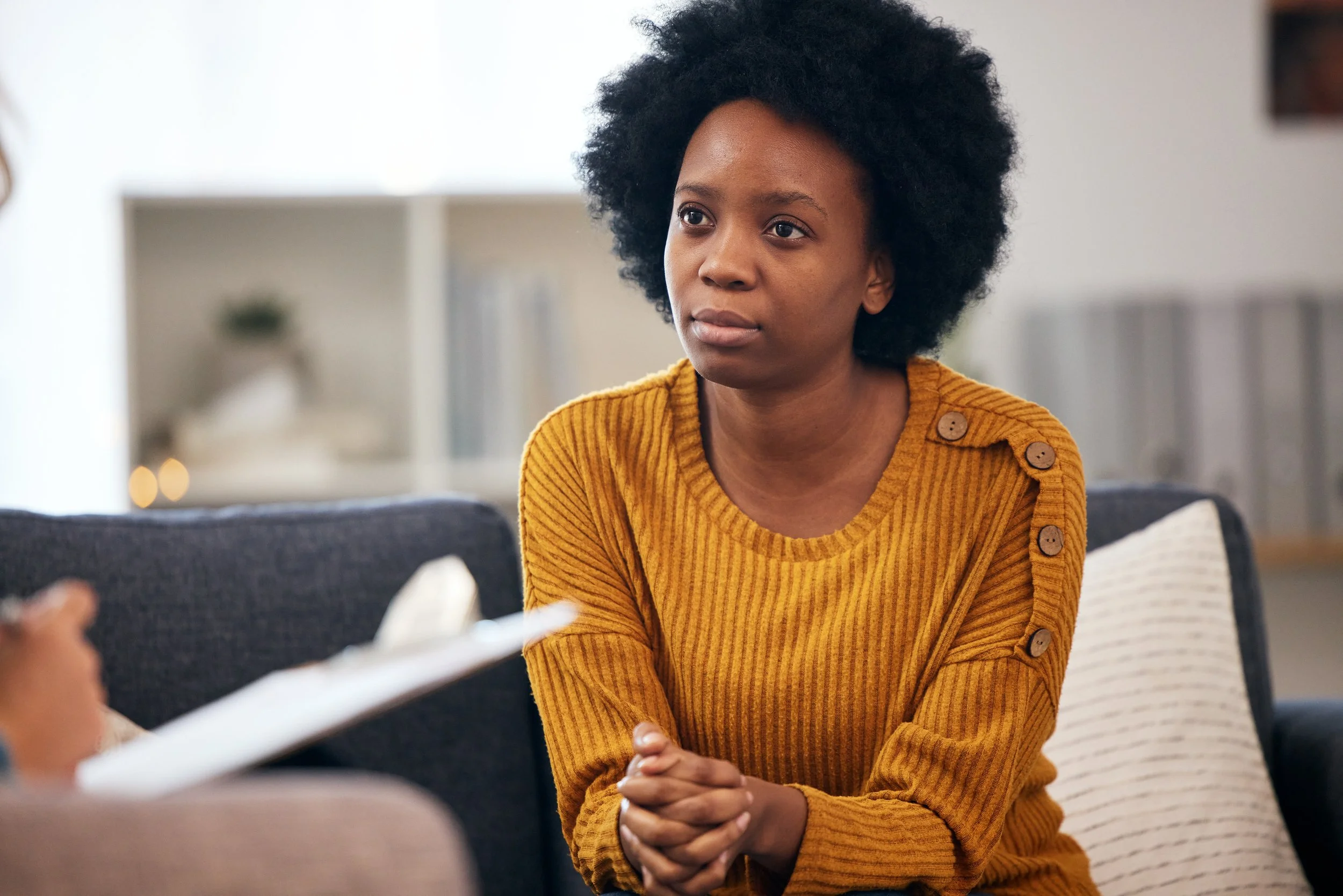 A woman with natural curly hair wearing a mustard yellow sweater with button details on the shoulder, sitting on a couch and engaging in conversation with someone out of the frame.