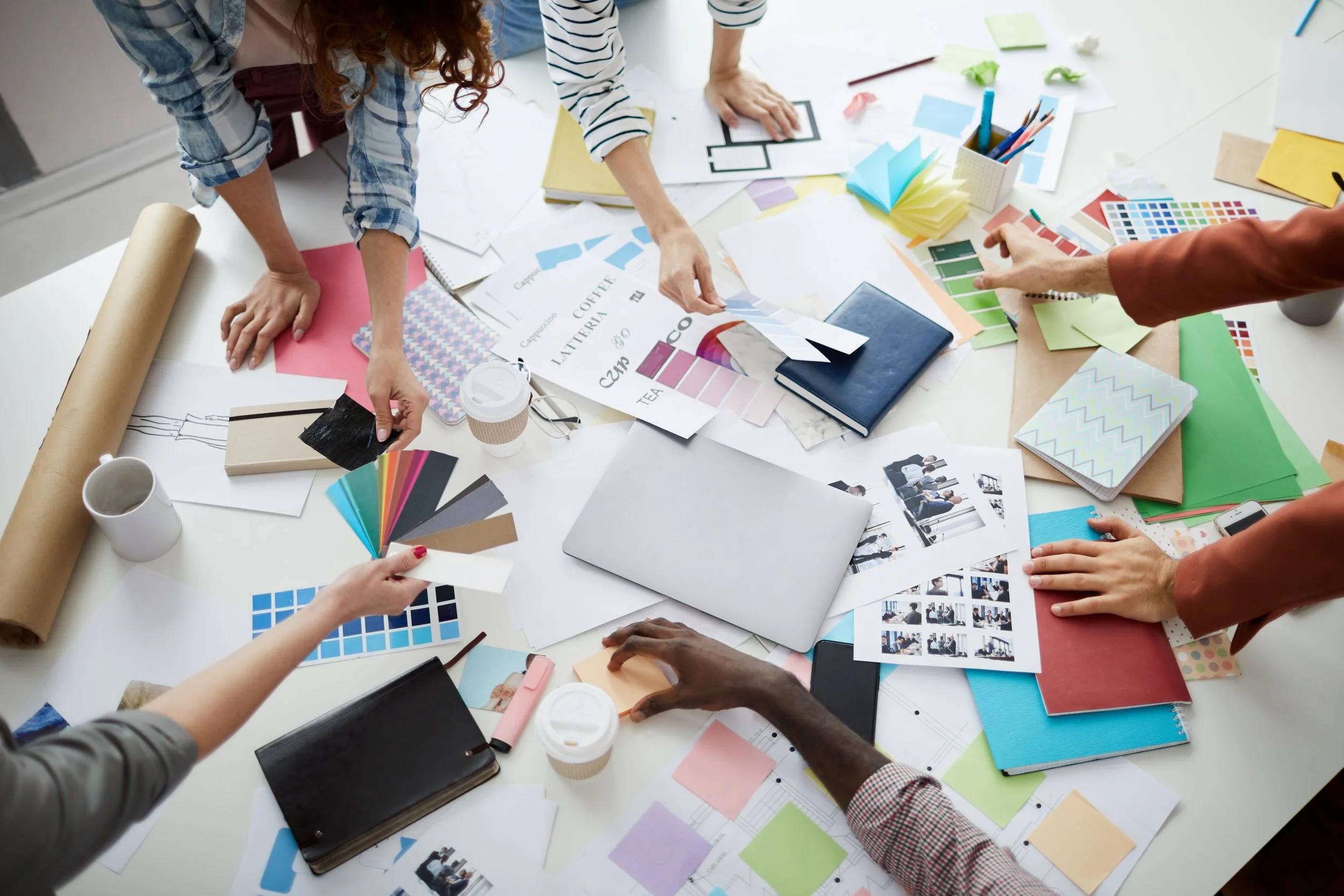 People working at a cluttered table covered with color swatches, papers, notebooks, and design materials.