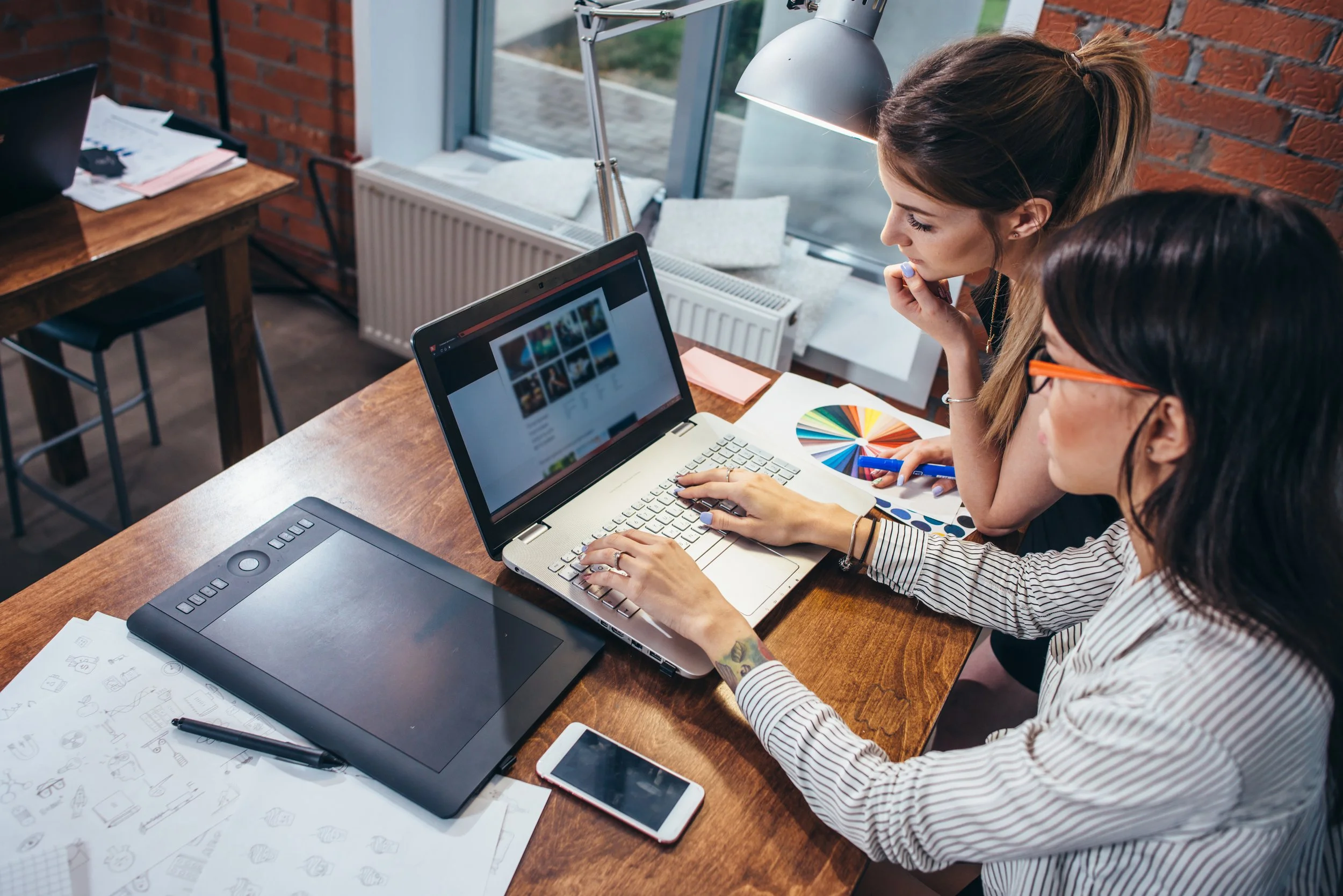 Two women working at a wooden desk with a laptop, a drawing tablet, and various design sketches in a bright room with large windows.