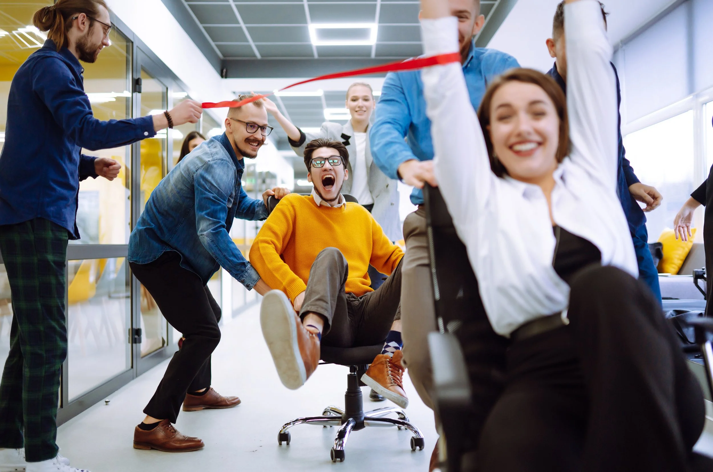 A group of young adults smiling and having fun in an office space. One person is seated in a chair while others stand around, some playfully pulling an office chair rope. The scene captures a joyful, lively moment among friends or colleagues.