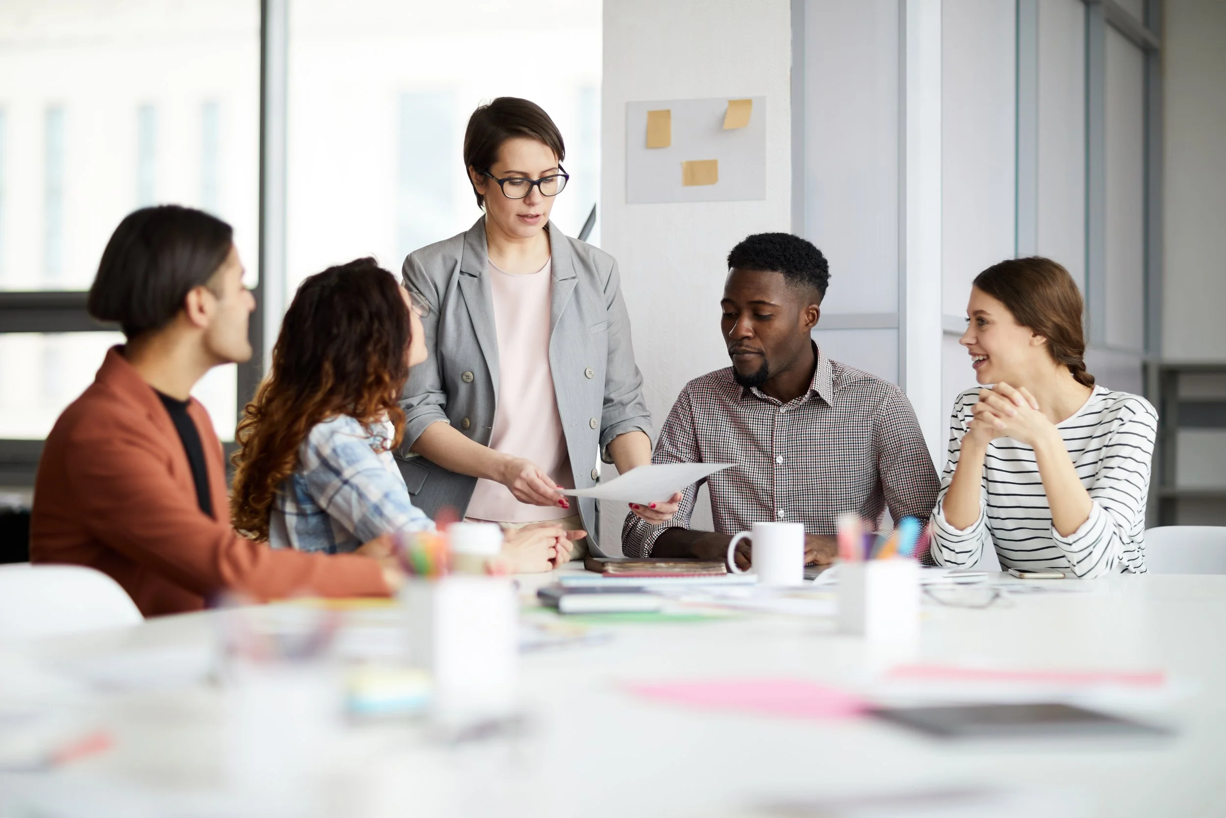 A diverse group of five people gathered around a table during a meeting, with a woman in glasses standing and holding papers while speaking to the group inside a bright office space.