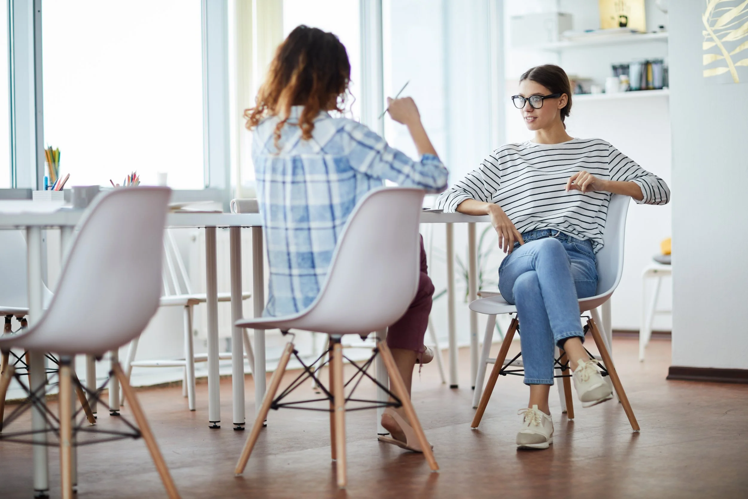 Two women are sitting in an office or conference room, engaged in a conversation. One has curly hair and is wearing a plaid shirt, while the other has straight hair, glasses, and is wearing a striped shirt. There are chairs and a table with colored pens and office supplies around them, with bright natural light coming through large windows.