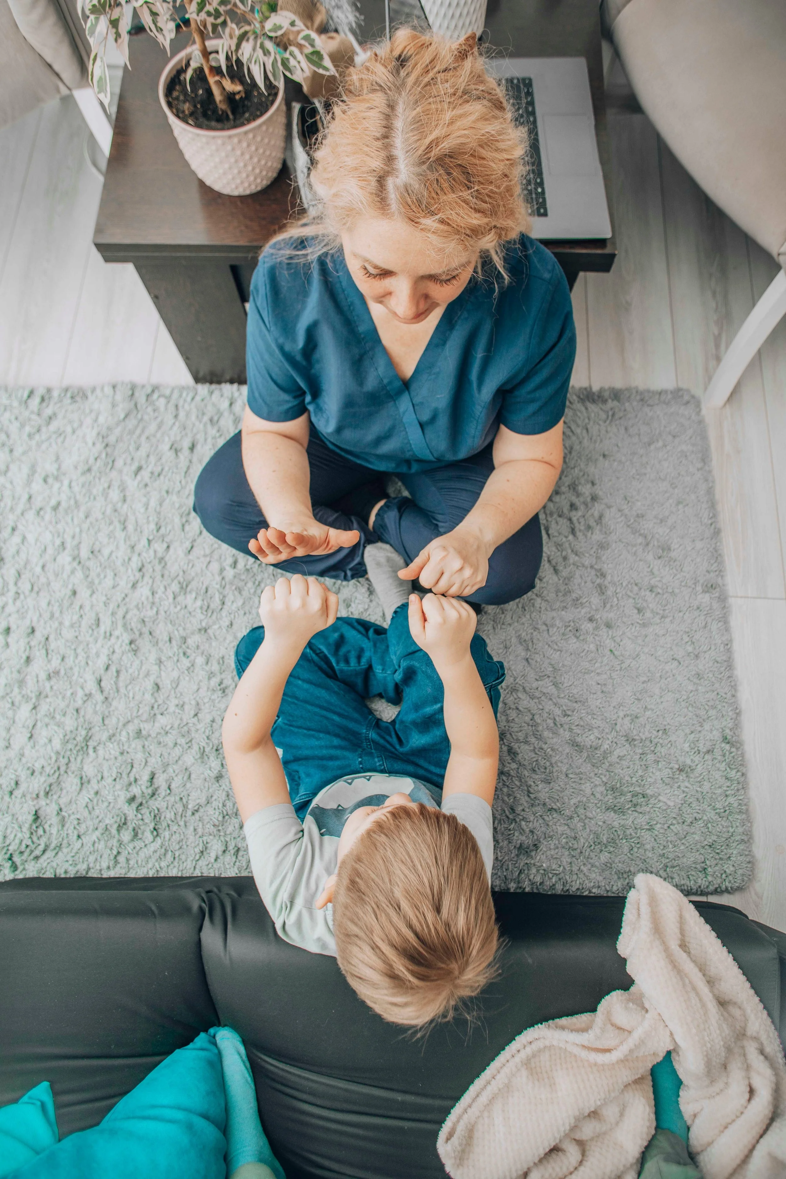 An adult woman with blonde hair and wearing blue scrubs interacts with a young boy sitting on a black leather couch. The woman is sitting on a gray rug with a coffee table behind her, which holds a potted plant and a closed laptop. The boy has blonde hair, is wearing a gray shirt with an animal design, and is holding the woman's hands. In the foreground, a teal blanket and a cream-colored blanket are visible.