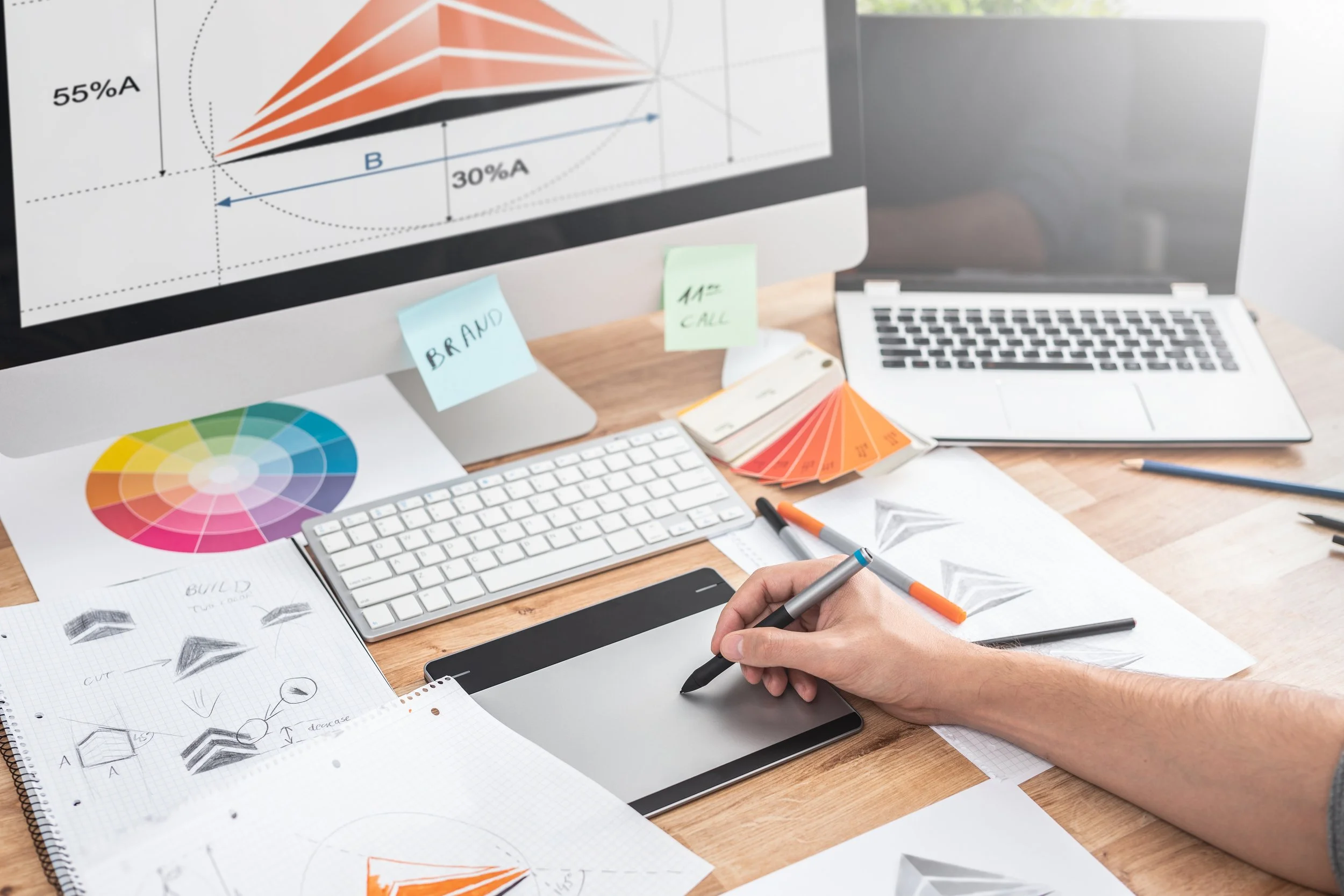 A workspace with color swatches, sketches, and stationery, a large monitor displaying a graph, a keyboard, a laptop, and various pens and papers on a wooden desk.
