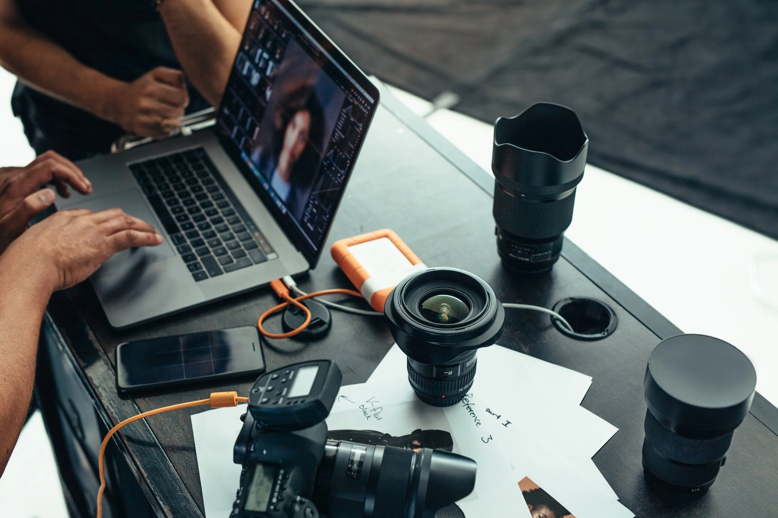 Photography workspace with camera lenses, a laptop showing photo editing software, a smartphone, handwritten notes, and photography equipment on a black table.