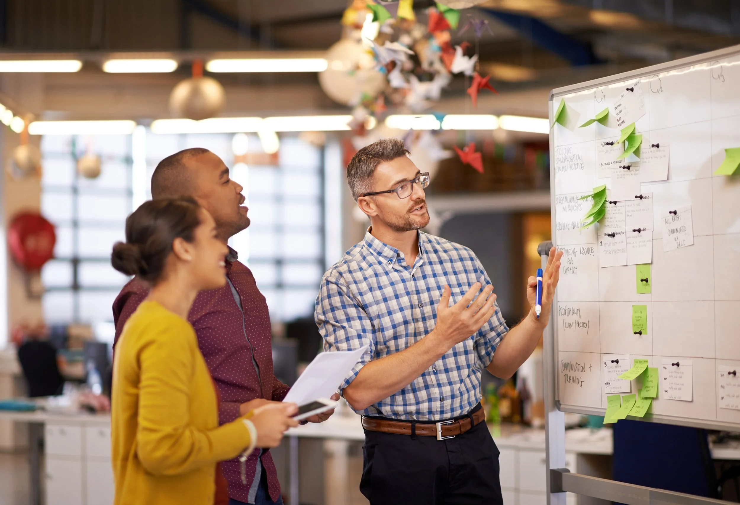 Three people in a meeting room looking at a whiteboard filled with sticky notes and diagrams, engaged in a discussion.