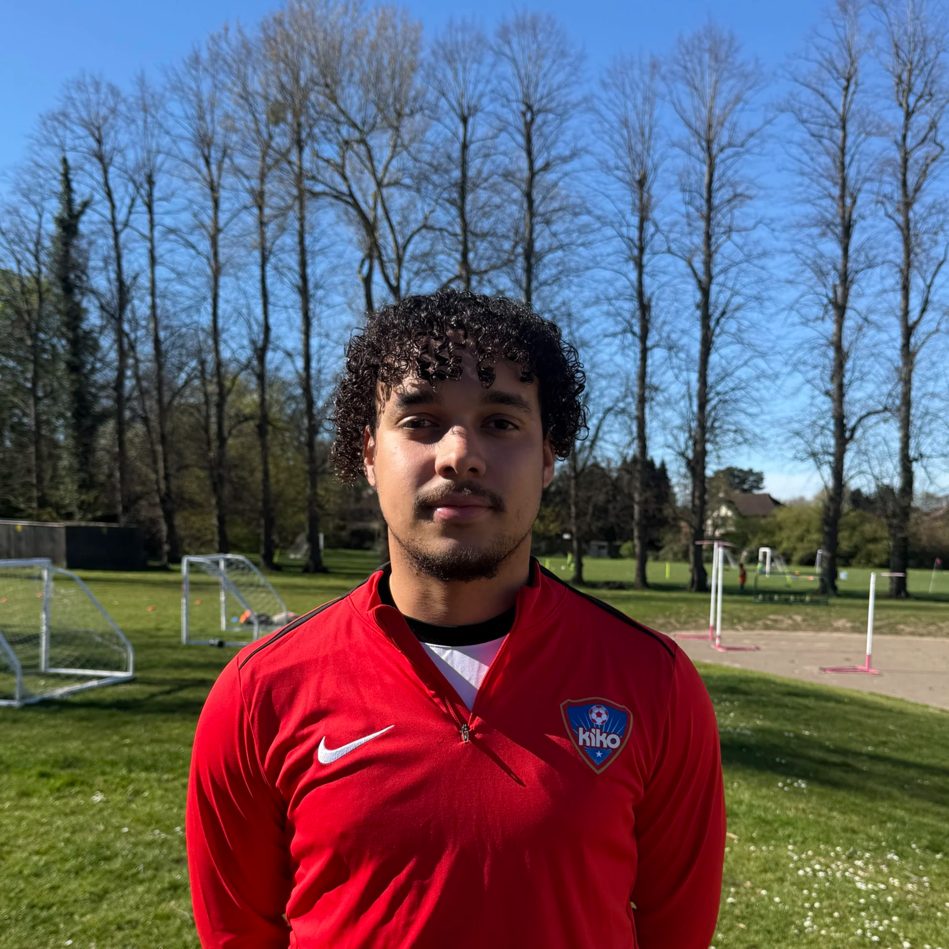 Young man with curly black hair and light brown skin wearing a red sports jacket with a club crest, standing outdoors in a park with leafless trees and agility training hurdles in the background on a sunny day.