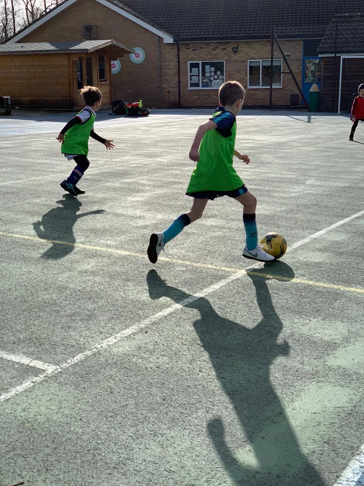 Children playing footballon an outdoor court, two wearing green vests, one dribbling the ball.