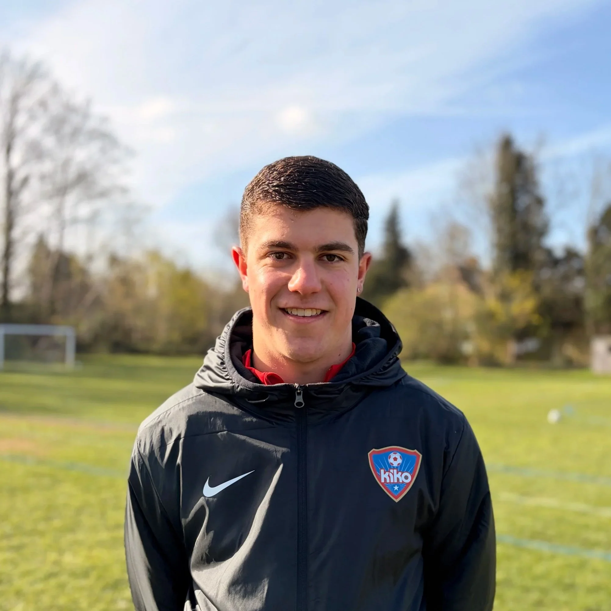 Close-up of a young man with dark hair smiling outdoors on a sunny day, wearing a black Nike sports jacket with a red and blue logo.