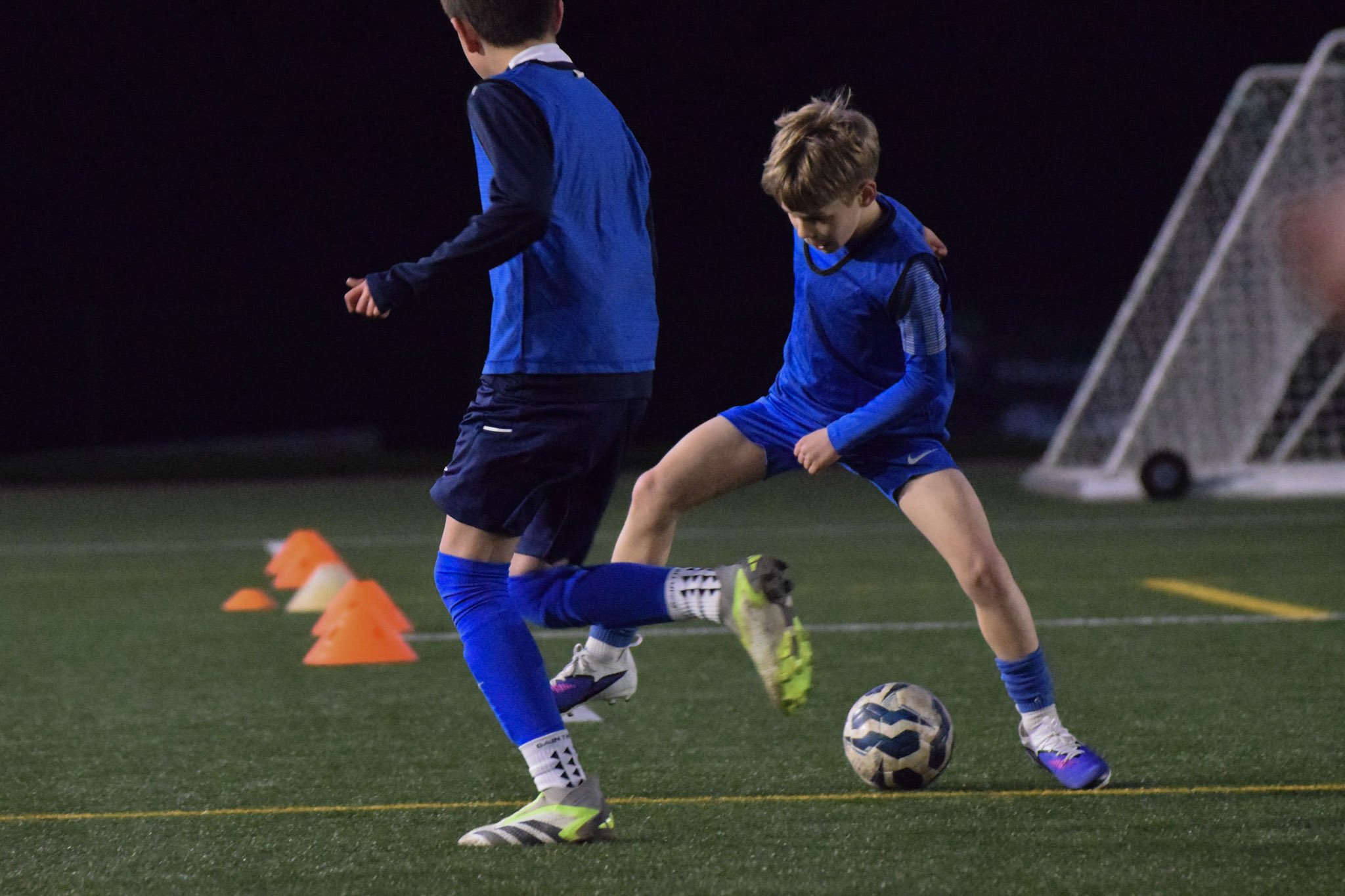 A young boy in a red soccer jersey and blue shorts running on a grassy soccer field, about to kick a soccer ball, with soccer goals and other people in the background.