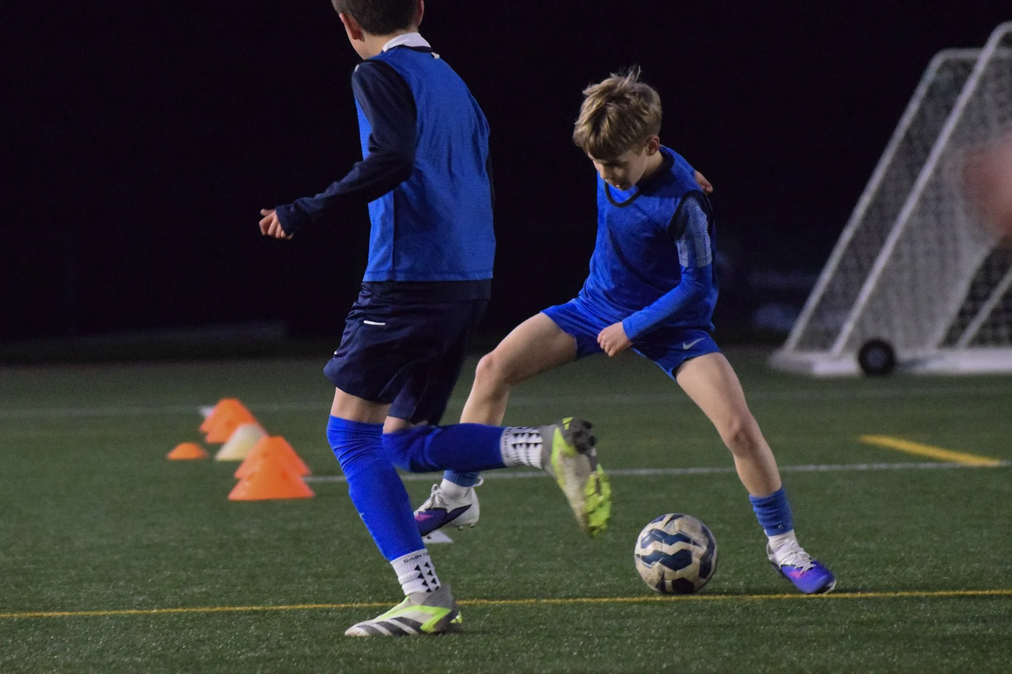 A young boy in a red soccer jersey and blue shorts running on a grassy soccer field, about to kick a soccer ball, with soccer goals and other people in the background.