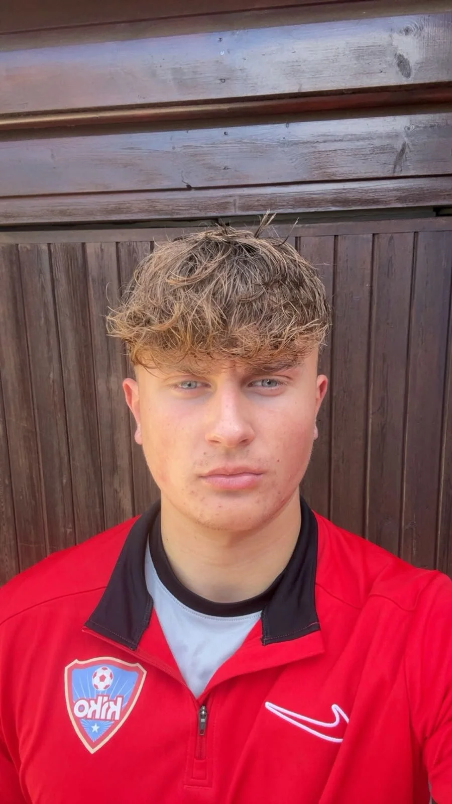 A young man with blond, wavy hair and blue eyes taking a selfie in front of a wooden fence. He's wearing a red sports jacket with a logo on the left side that features a soccer ball and the word 'OHIO'.