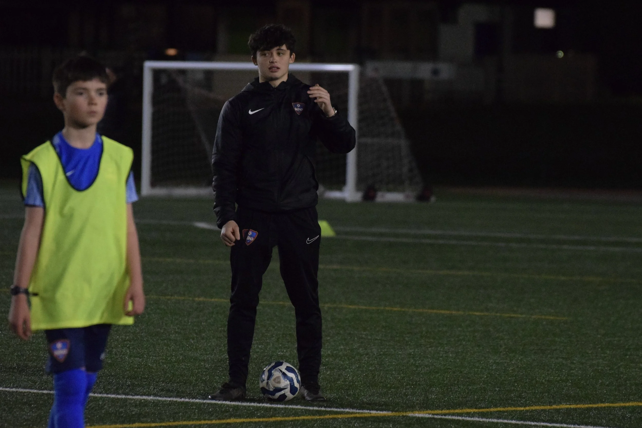 A young man in sportswear standing on a soccer field at night with a soccer ball at his feet, and another kid in a yellow vest visible in the foreground.