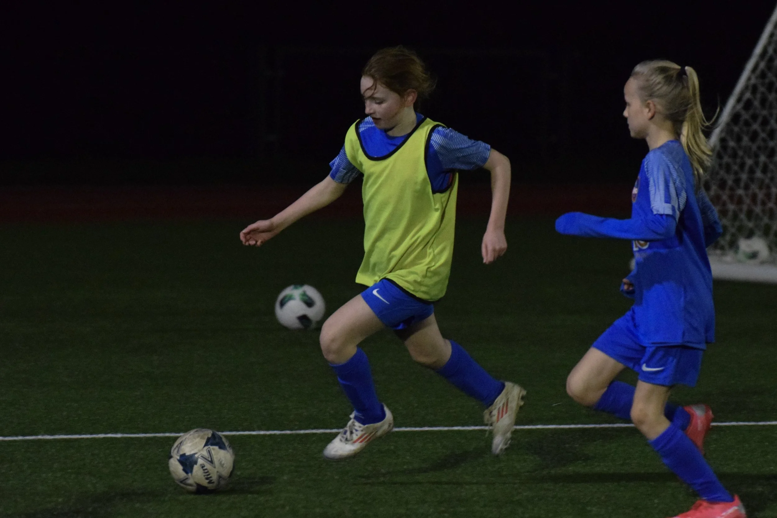 Two young girls playing soccer at night, one wearing a yellow vest and the other in a blue jersey, chasing a soccer ball on a dark field with a goalpost in the background.