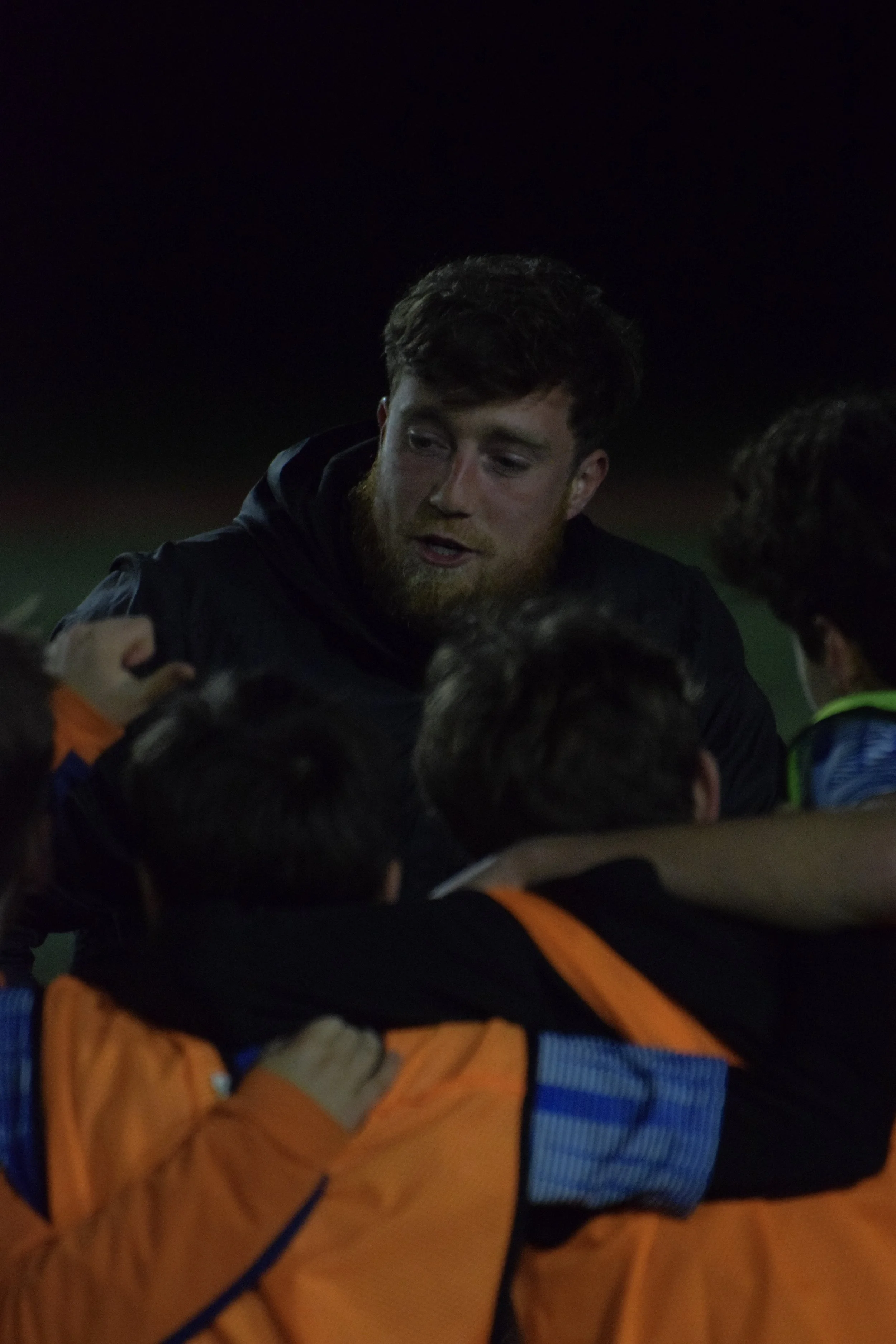 A group of people hugging outdoors at night, one man with brown hair and a beard in the center, wearing a black jacket.
