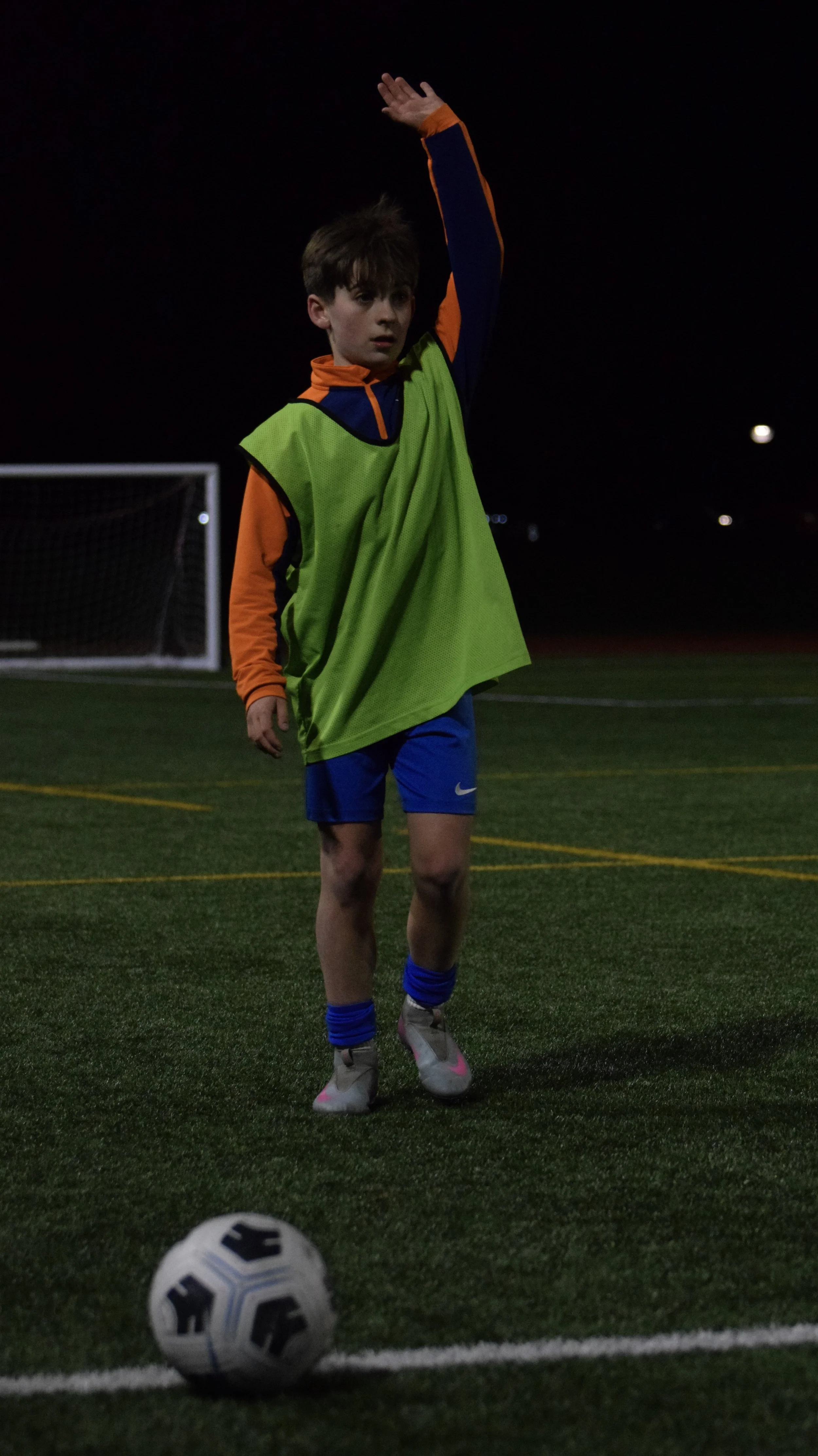 A young boy in sports attire about to kick a soccer ball on a soccer field at night, with goalpost in the background.
