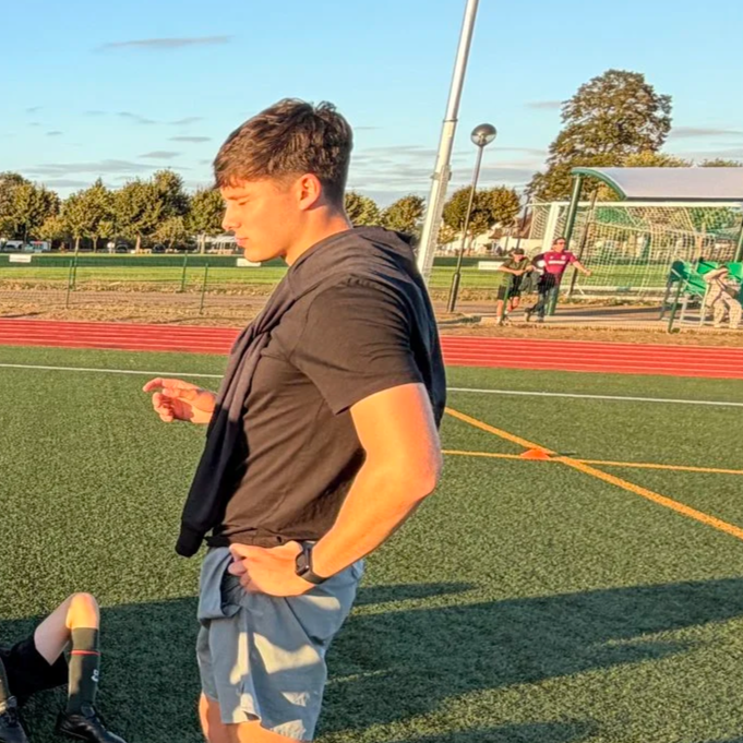 Young man standing on a sports field, looking at his phone, wearing a black T-shirt, gray shorts, and a watch, with a soccer goal and people in the background.