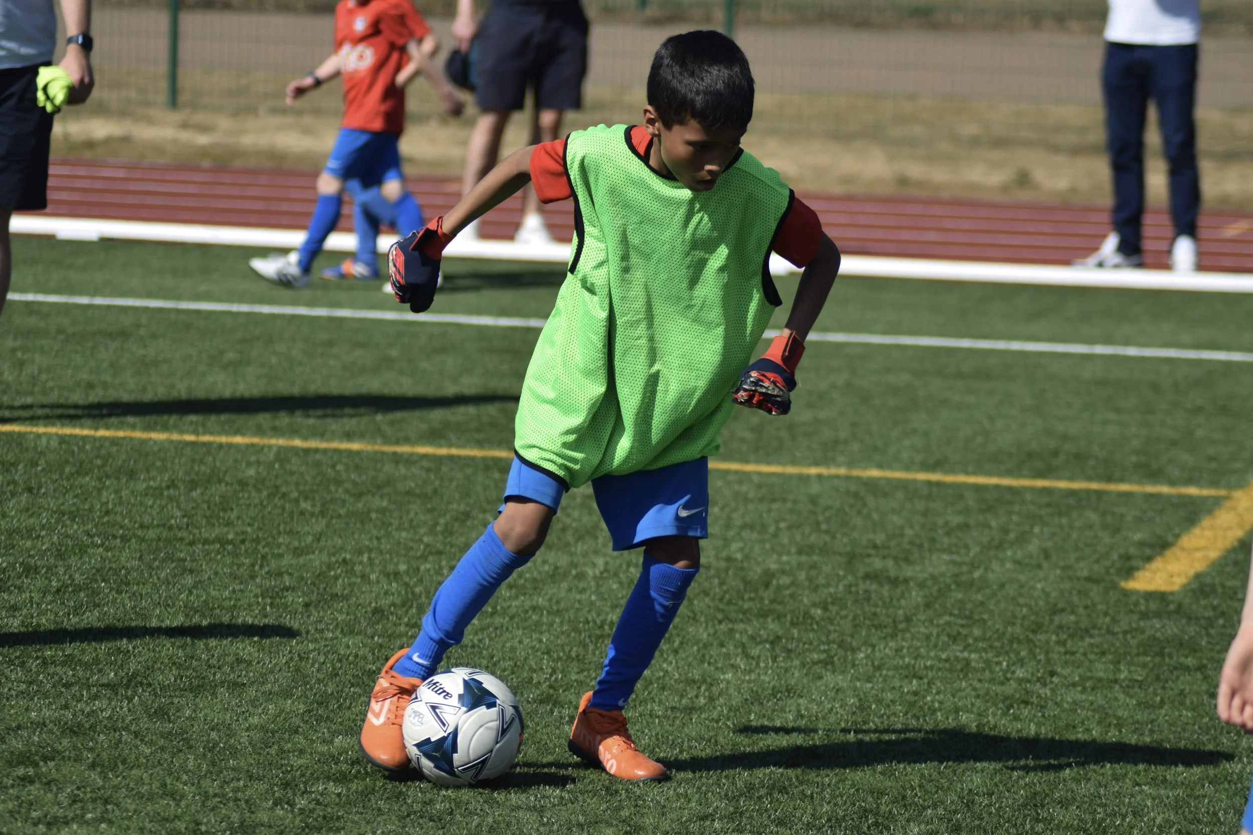 A young boy in a red shirt, blue shorts, and orange cleats is dribbling a soccer ball on a field, wearing a green training vest.
